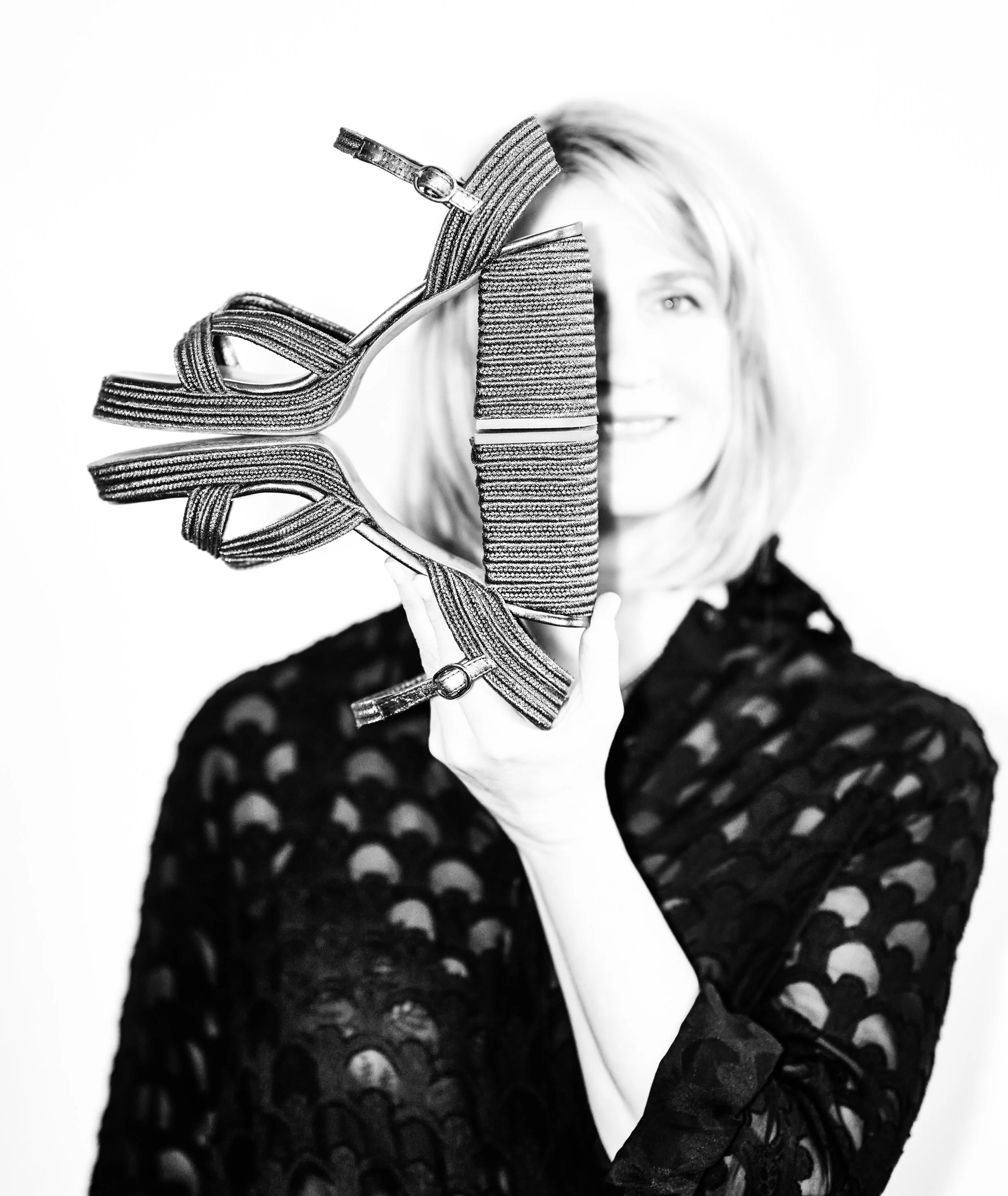 Black and white photo of a woman with wavy hair, holding a high-heeled shoe in front of her face. The shoe has multiple straps and a chunky heel. She is wearing a dark, lace-patterned dress.