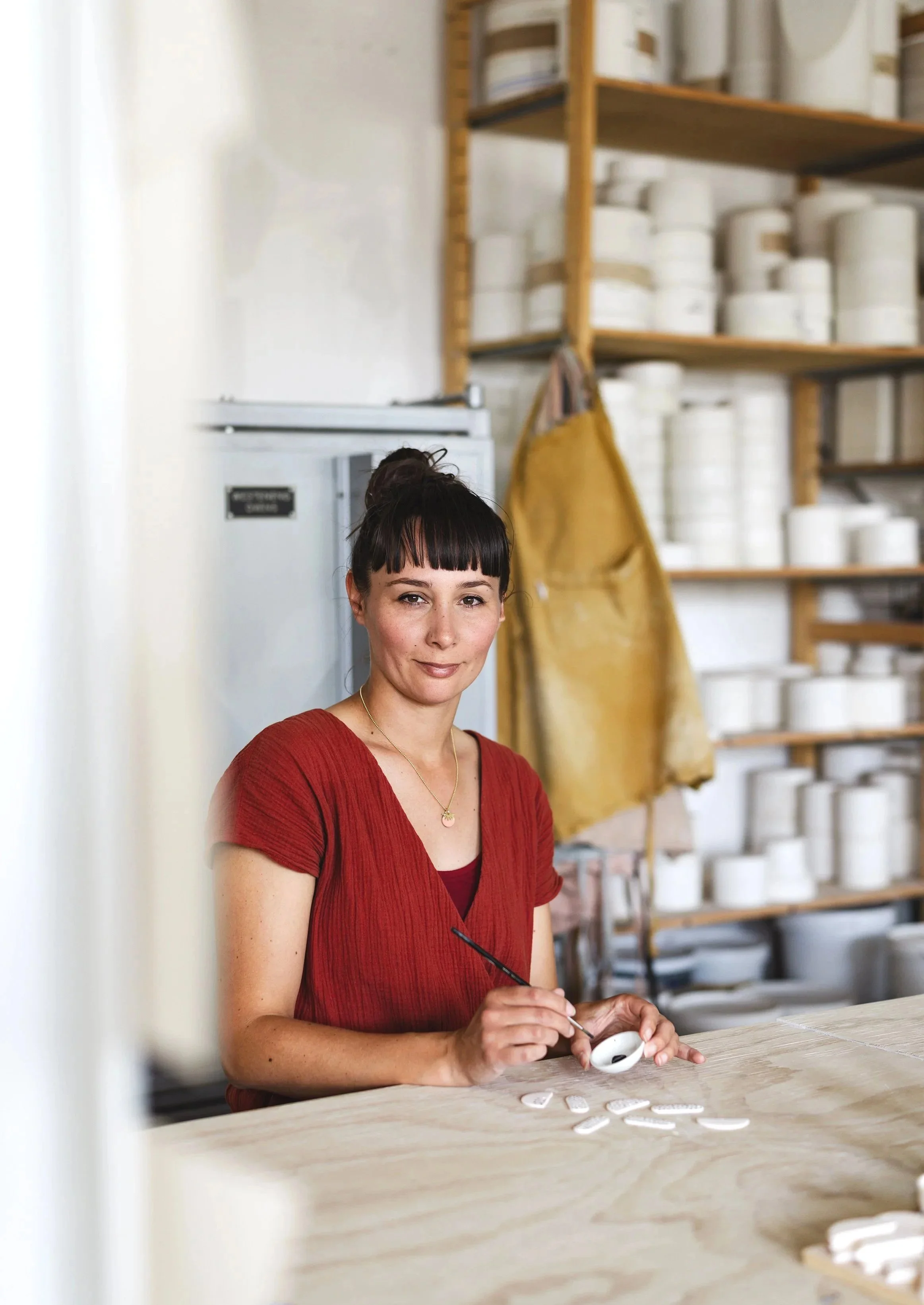 A woman with dark hair tied in a bun, wearing a red blouse, sits at a table in a pottery workshop, holding a paintbrush and painting a small ceramic piece.