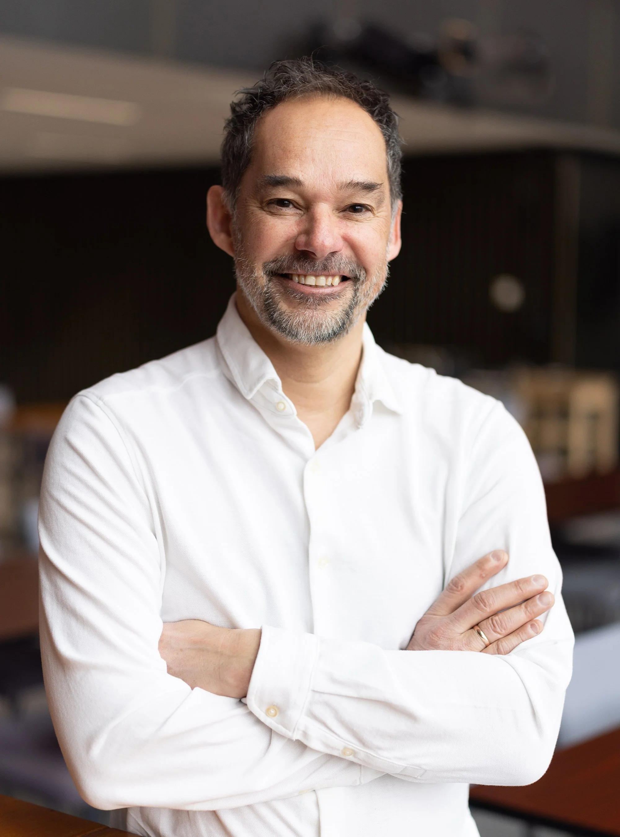 Smiling middle-aged man with a beard and mustache, wearing a white button-up shirt, standing with arms crossed in an indoor setting.