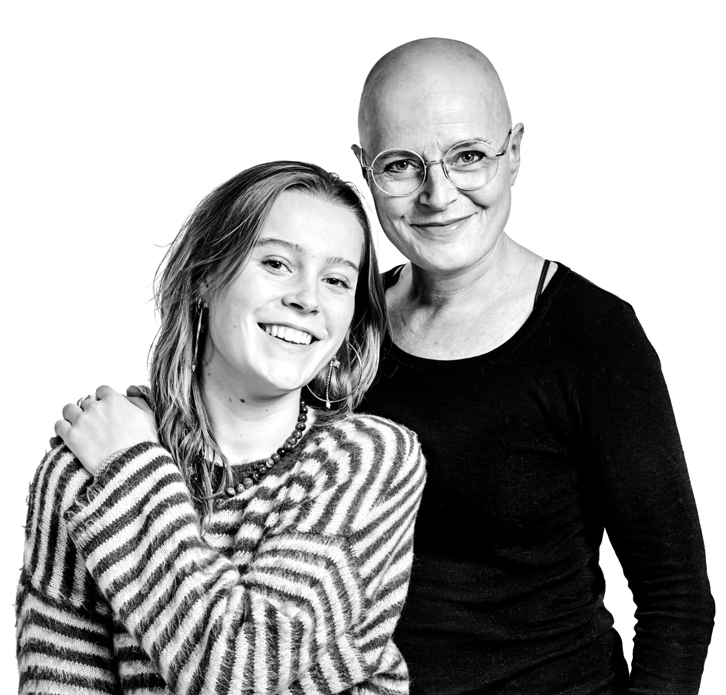 Black and white portrait of two smiling women, one young with shoulder-length hair wearing a striped sweater and jewelry, and the other older with a shaved head wearing glasses and a black top, posing together against a white background.