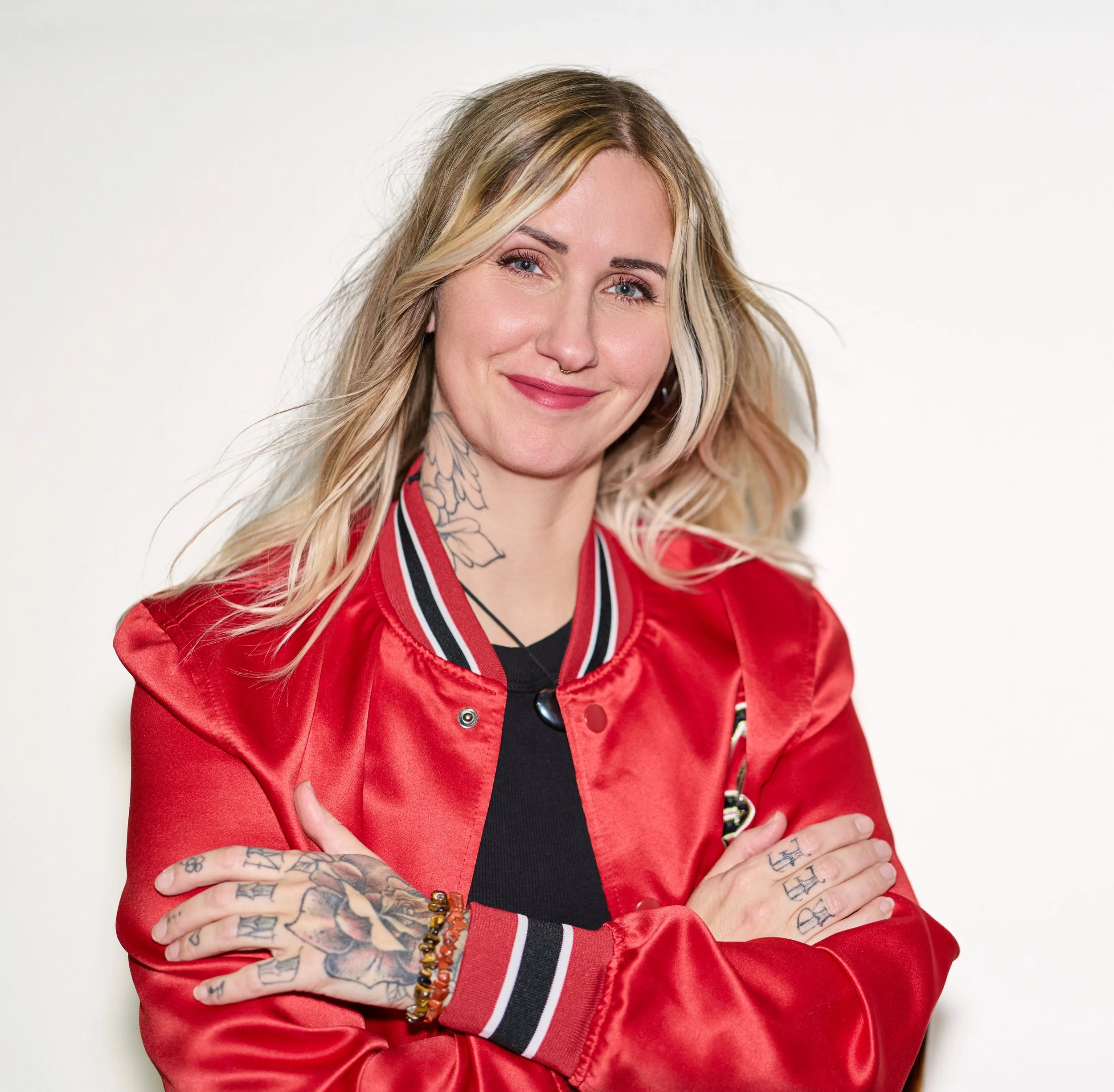 A smiling woman with tattoos on her hands and neck, blonde hair, wearing a red satin varsity jacket, standing against a plain white background.
