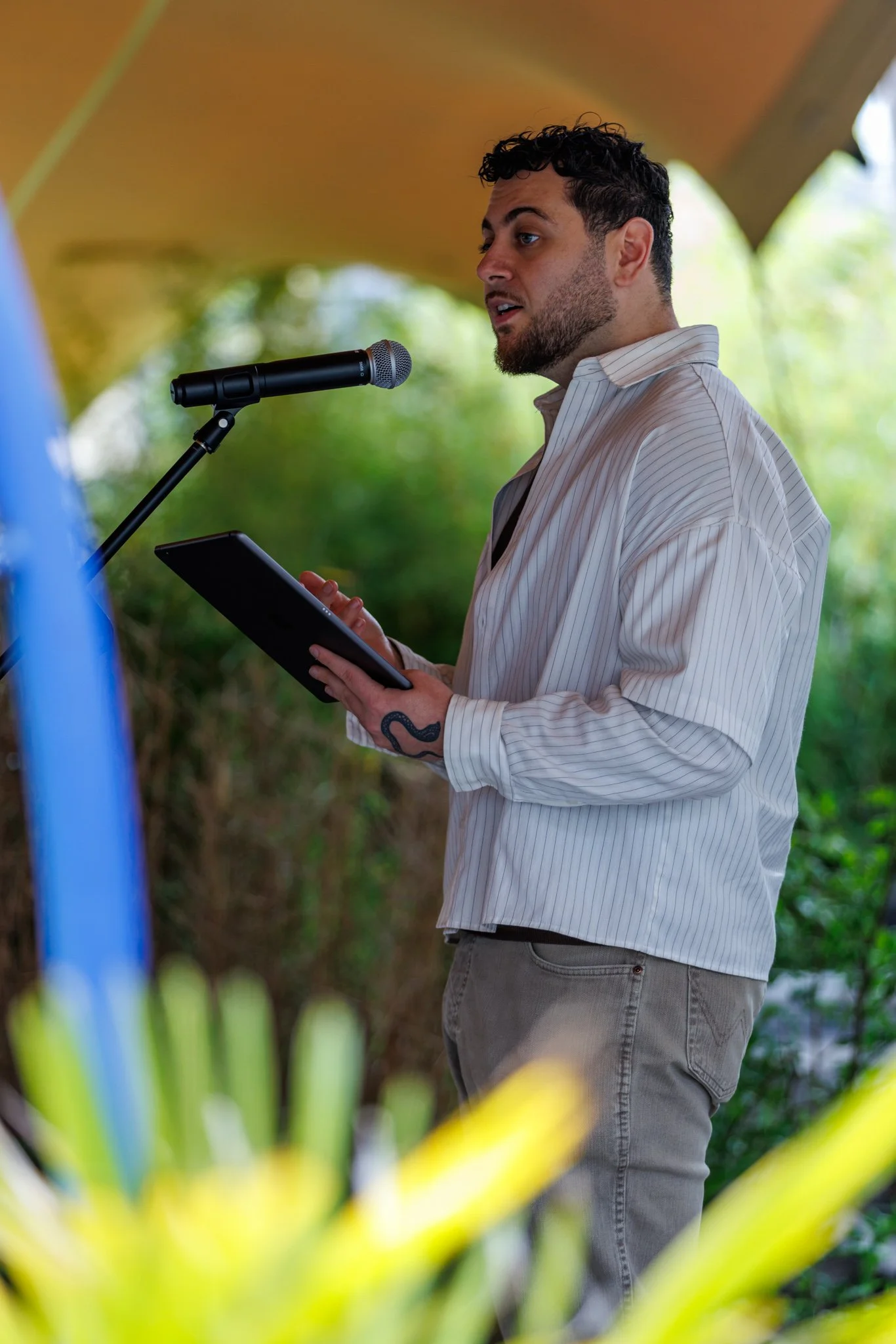 Man with dark curly hair and beard speaking into microphone while holding a tablet, outdoors with a blurred green background.