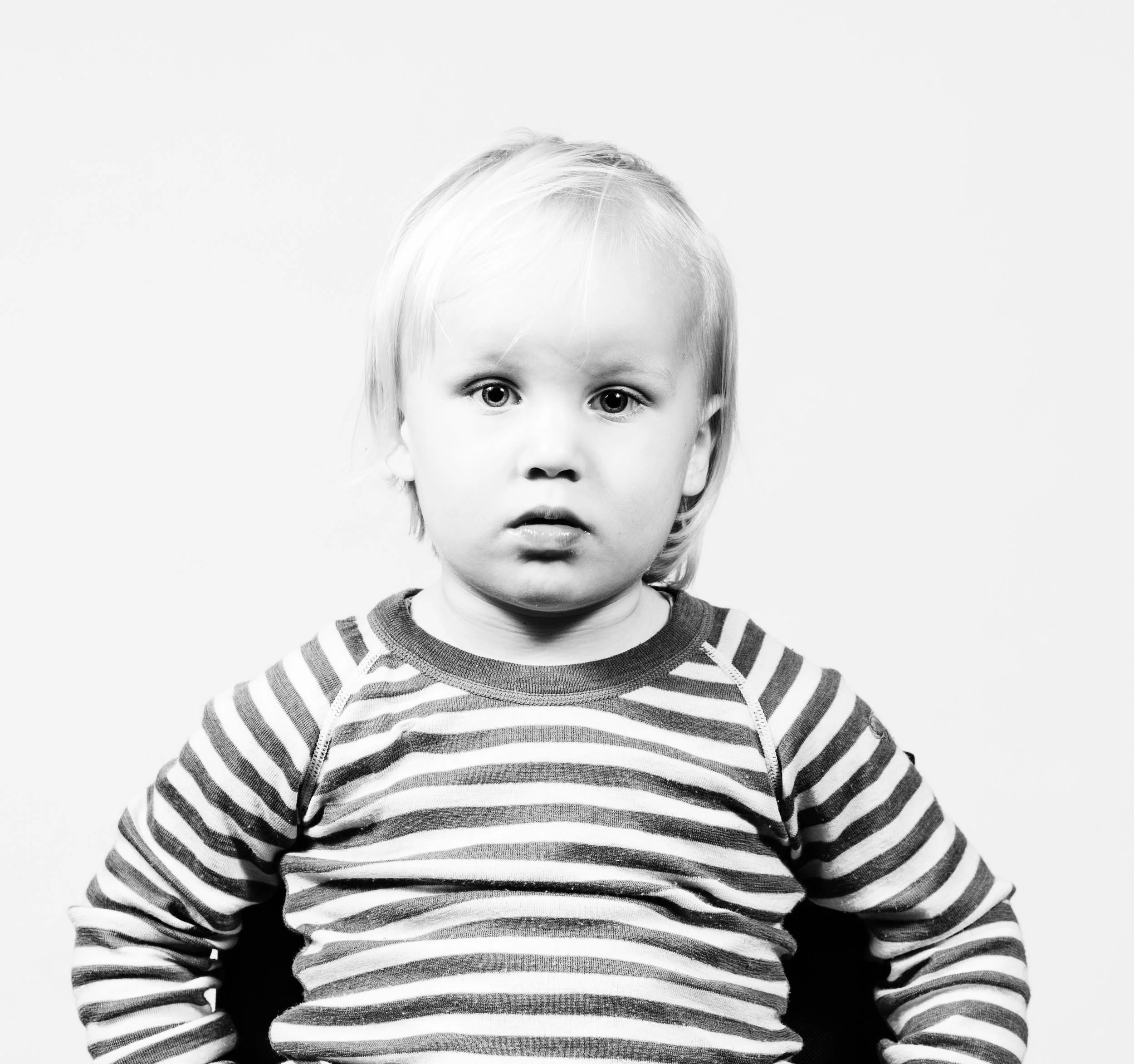 A young child with light hair and big eyes, wearing a striped long-sleeve shirt, looking directly at the camera with a serious expression.
