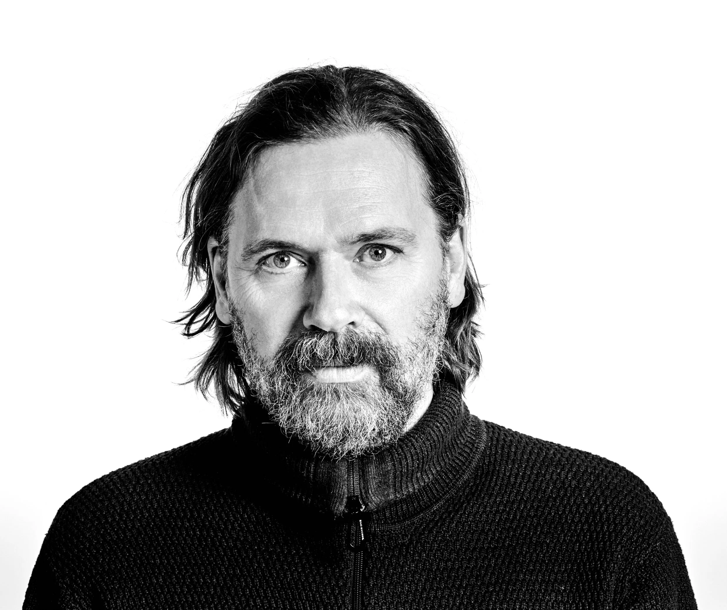 Black and white portrait of a middle-aged man with shoulder-length hair, beard, and mustache, wearing a textured zip-up sweater against a white background.