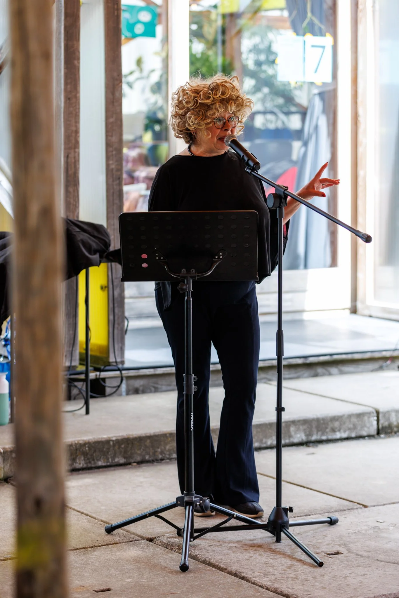 A woman with curly blonde hair wearing glasses and black clothing stands at a microphone stand, gesturing with her right hand while speaking or singing. She is positioned outdoors near a building with large glass windows and steps leading up to the e