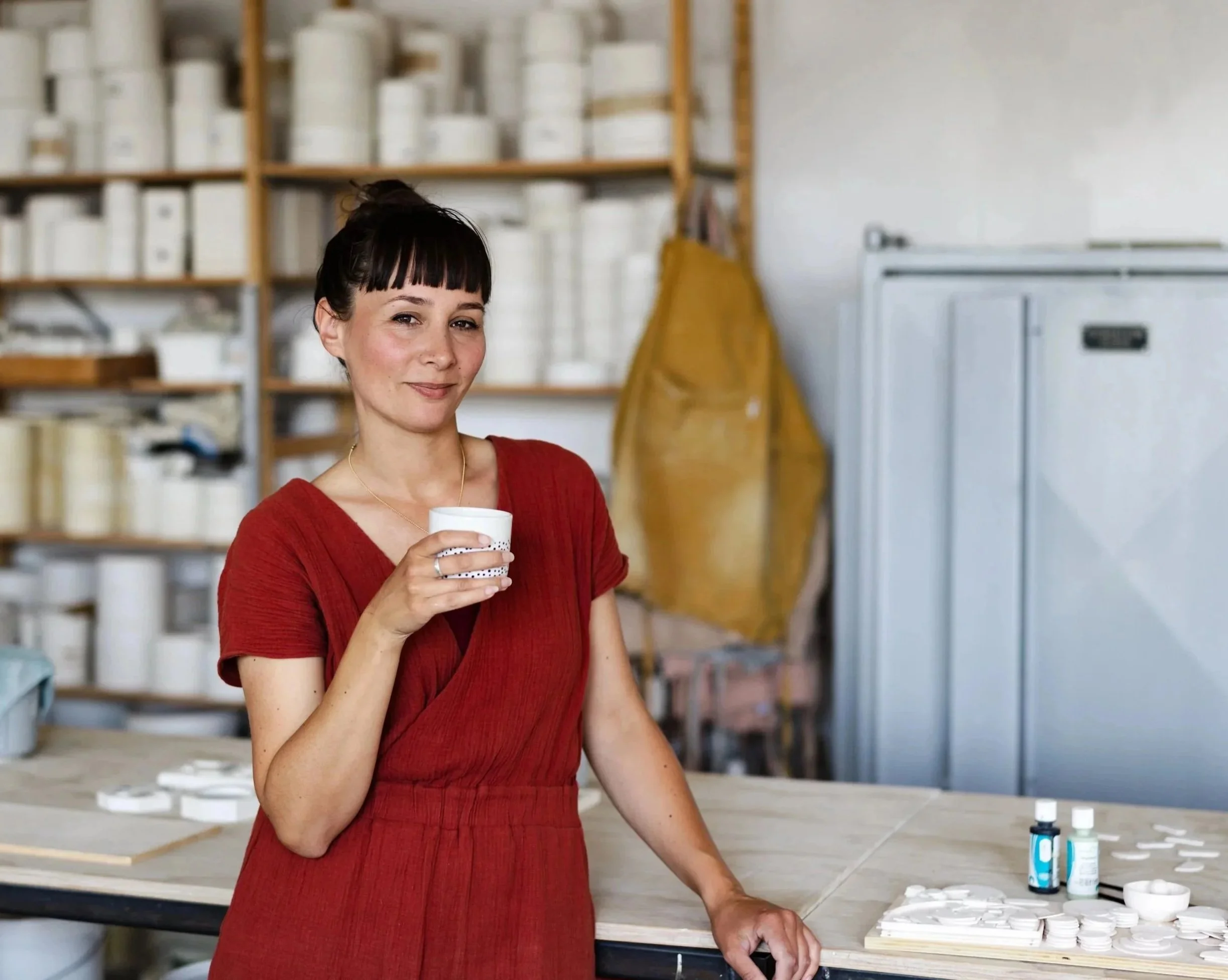 A woman in a red dress holding a white cup in a ceramics studio, with shelves of pottery and tools in the background.