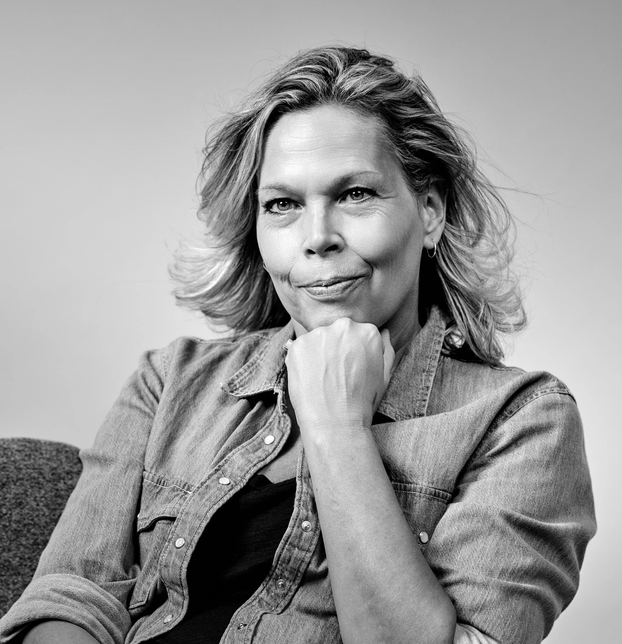 Vrouw met blond haar dat wappert door een windmachine, glimlacht naar de fotograaf terwijl ze met haar kin op een hand leunt