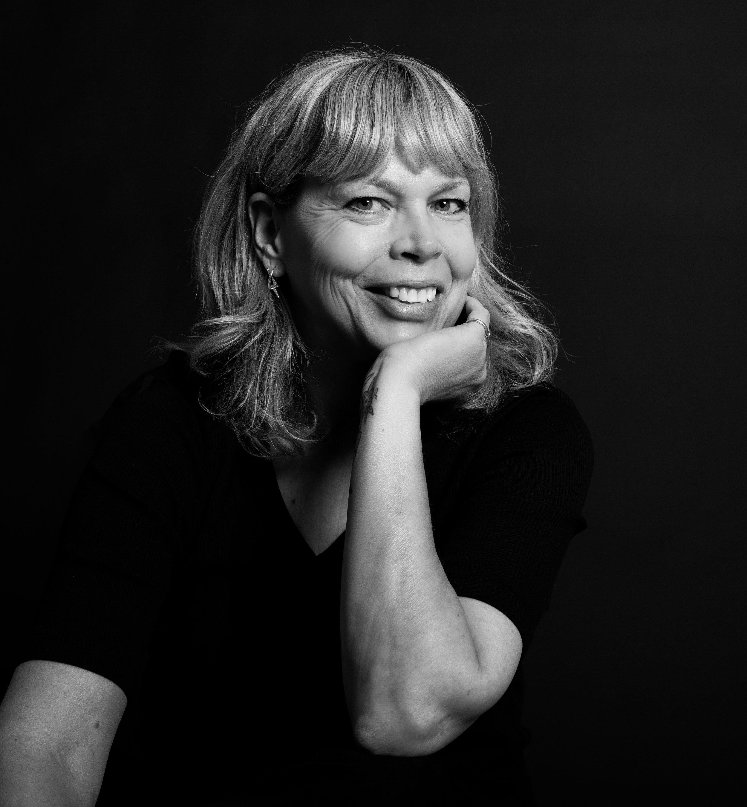 Black and white portrait of a smiling woman with shoulder-length wavy hair, resting her chin on her hand, against a dark background.