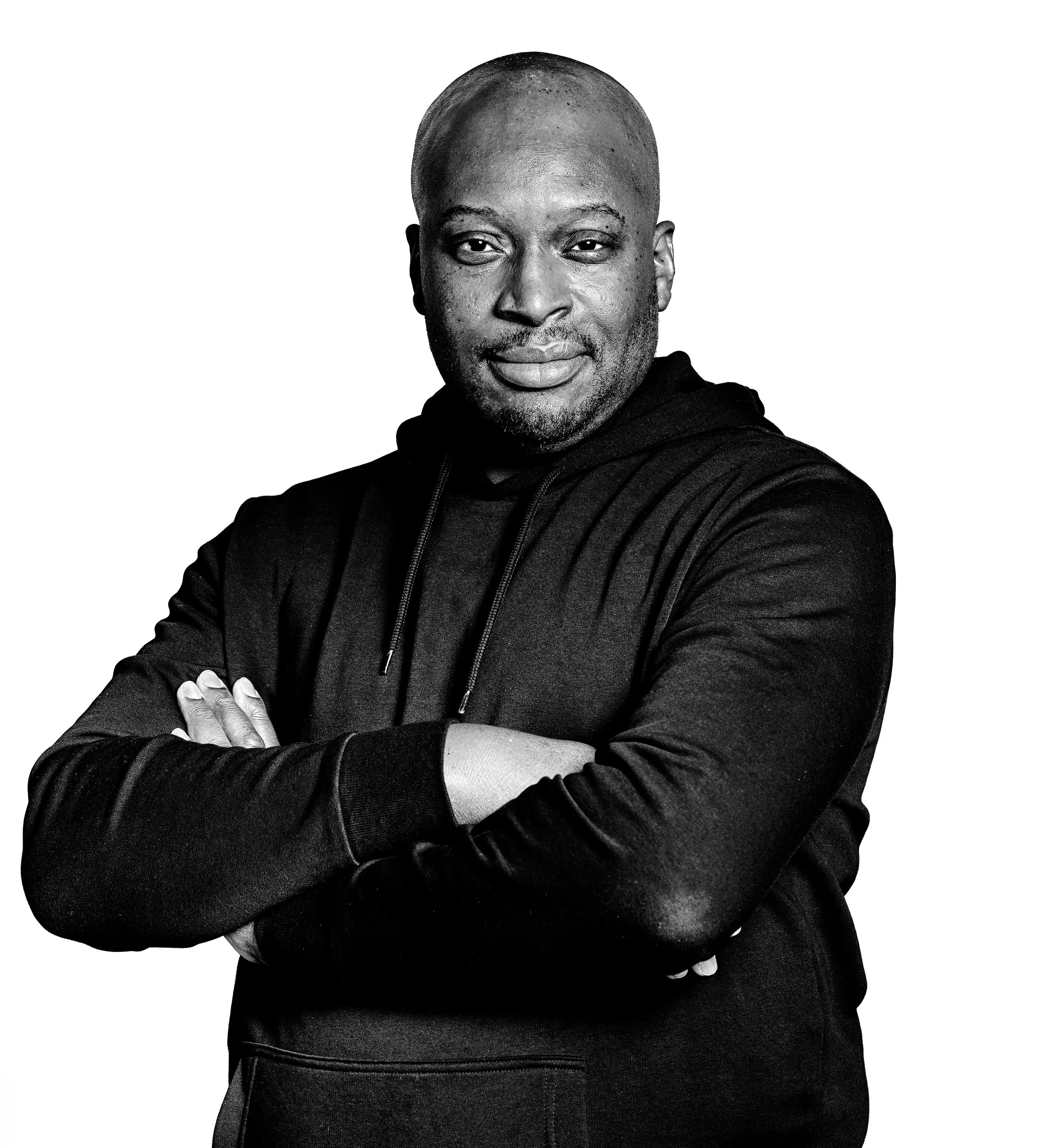 Black and white photo of a man with arms crossed, wearing a hoodie, smiling confidently.