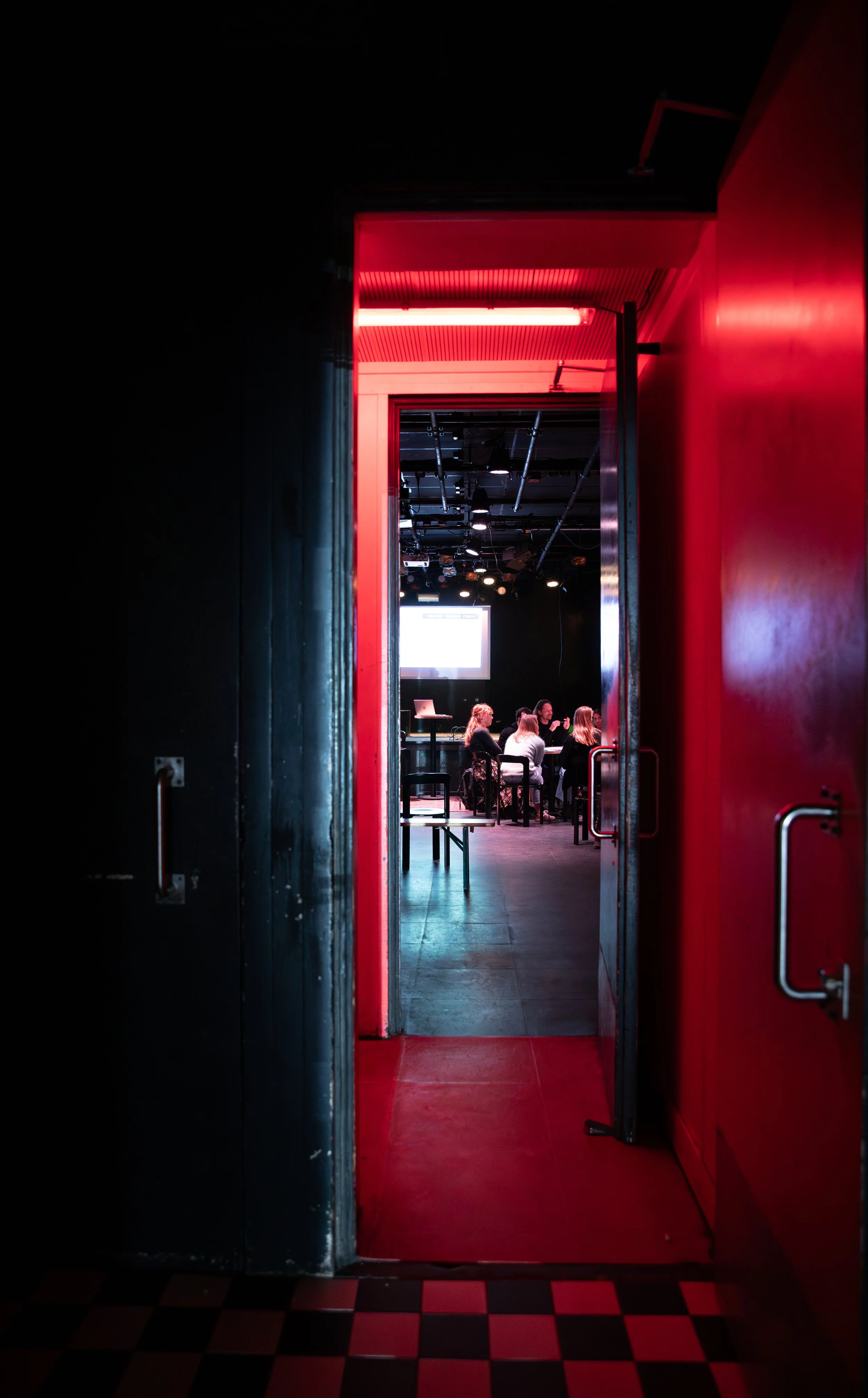 A doorway with red framing leads into a dark room where a small group of people are sitting at a table, with a projection screen visible in the background.