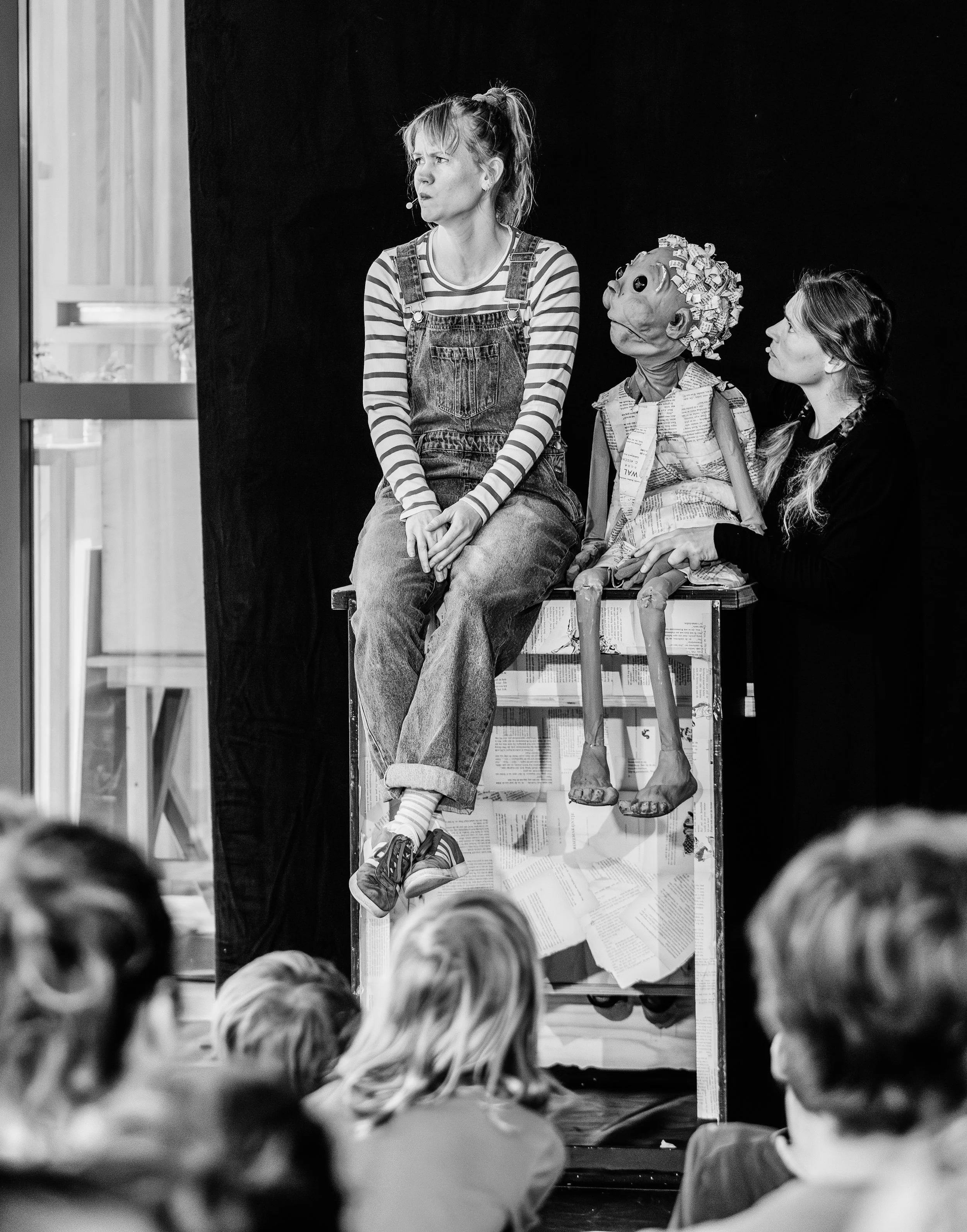 A woman with a microphone headset wearing striped shirt and overalls sits on a box, looking to her left, with a puppet with curly hair and large eyes, dressed in clothing made of newspaper, sitting beside her. The scene appears to be a puppet show or