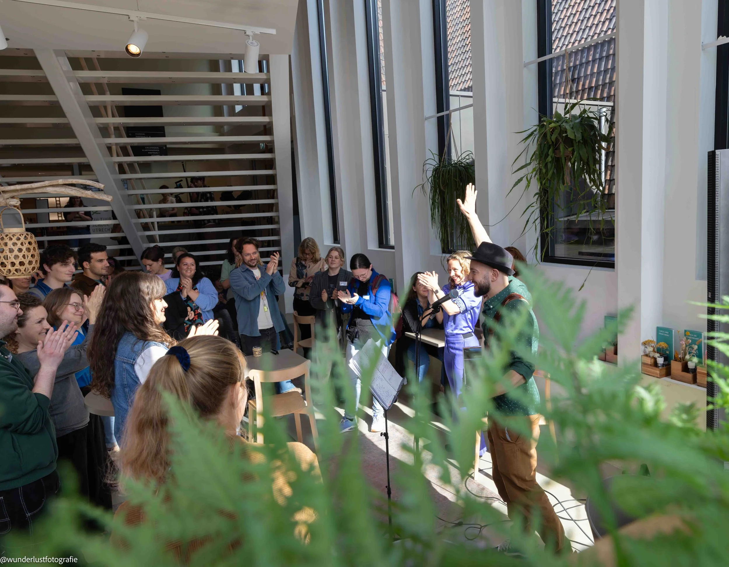 A group of people gathered indoors listening to a musician play guitar and sing into a microphone, with some clapping and smiling.
