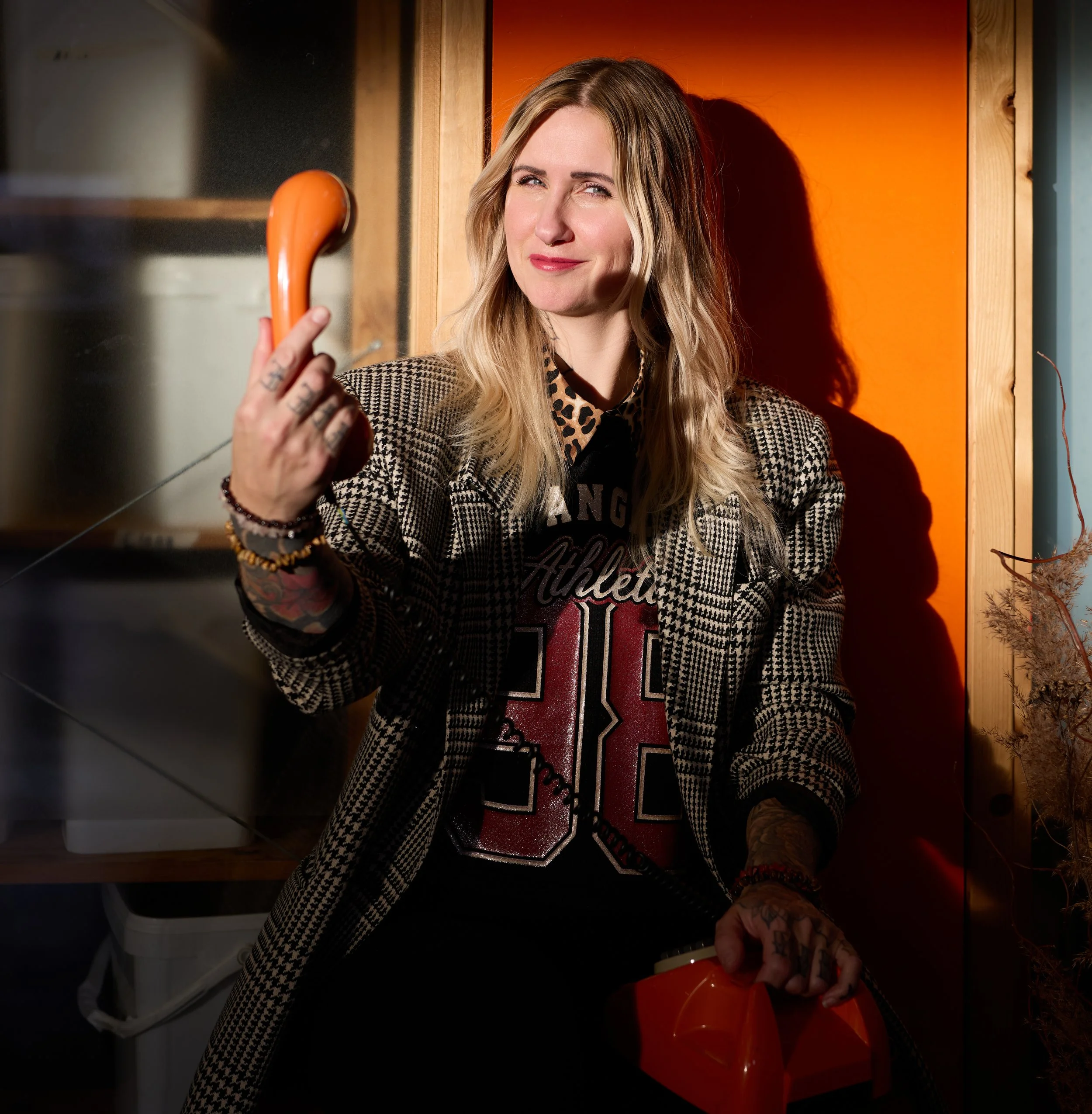 A woman with blonde hair wearing a checkered blazer and animal print shirt, holding an orange coffee mug, standing against an orange background with wooden trim.