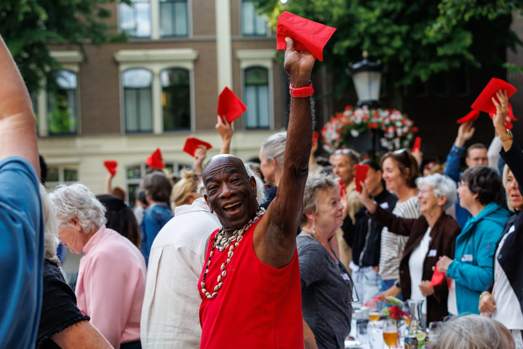 Een groep mensen die samen een feest of bijeenkomst vieren, met een man in een rode jurk en ketting die omhoog steekt, op een straat met huizen op de achtergrond.