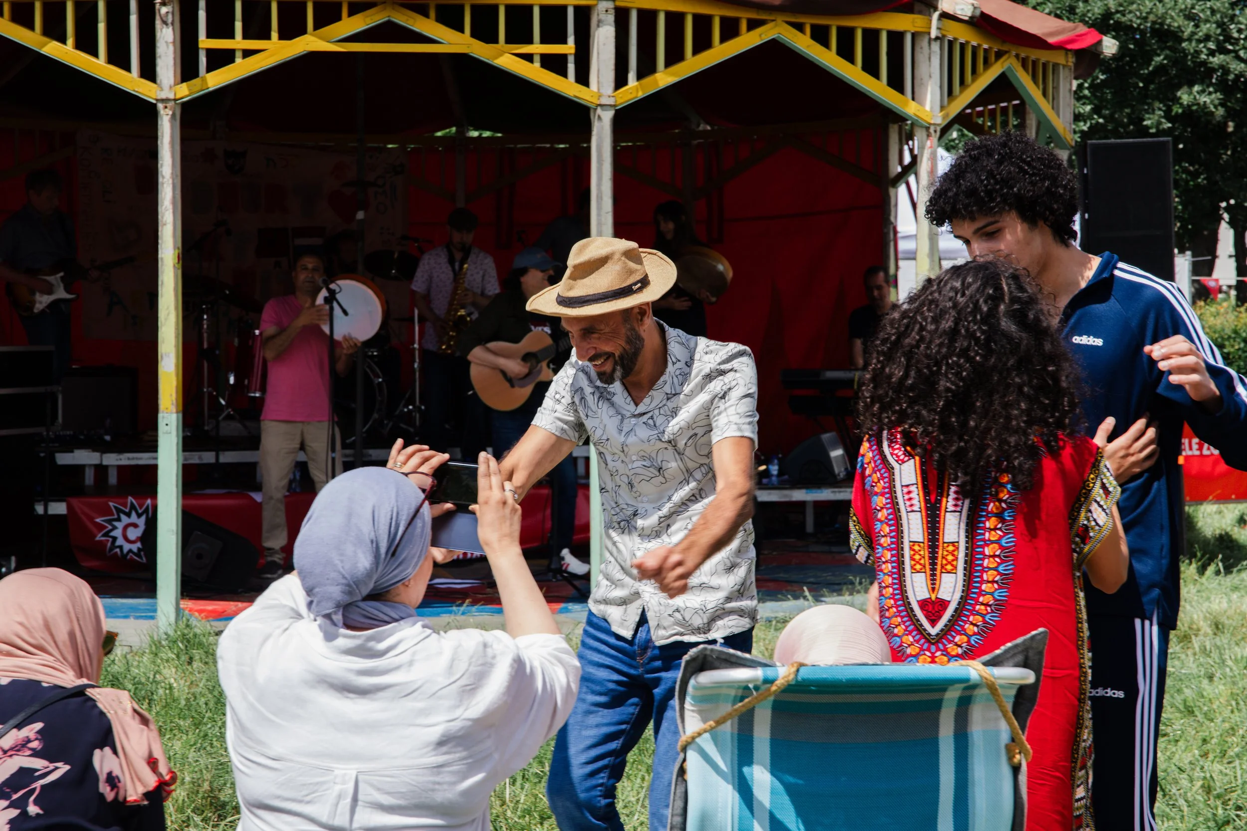 People dancing and smiling outdoors during a musical performance, with musicians playing instruments on a stage in the background.