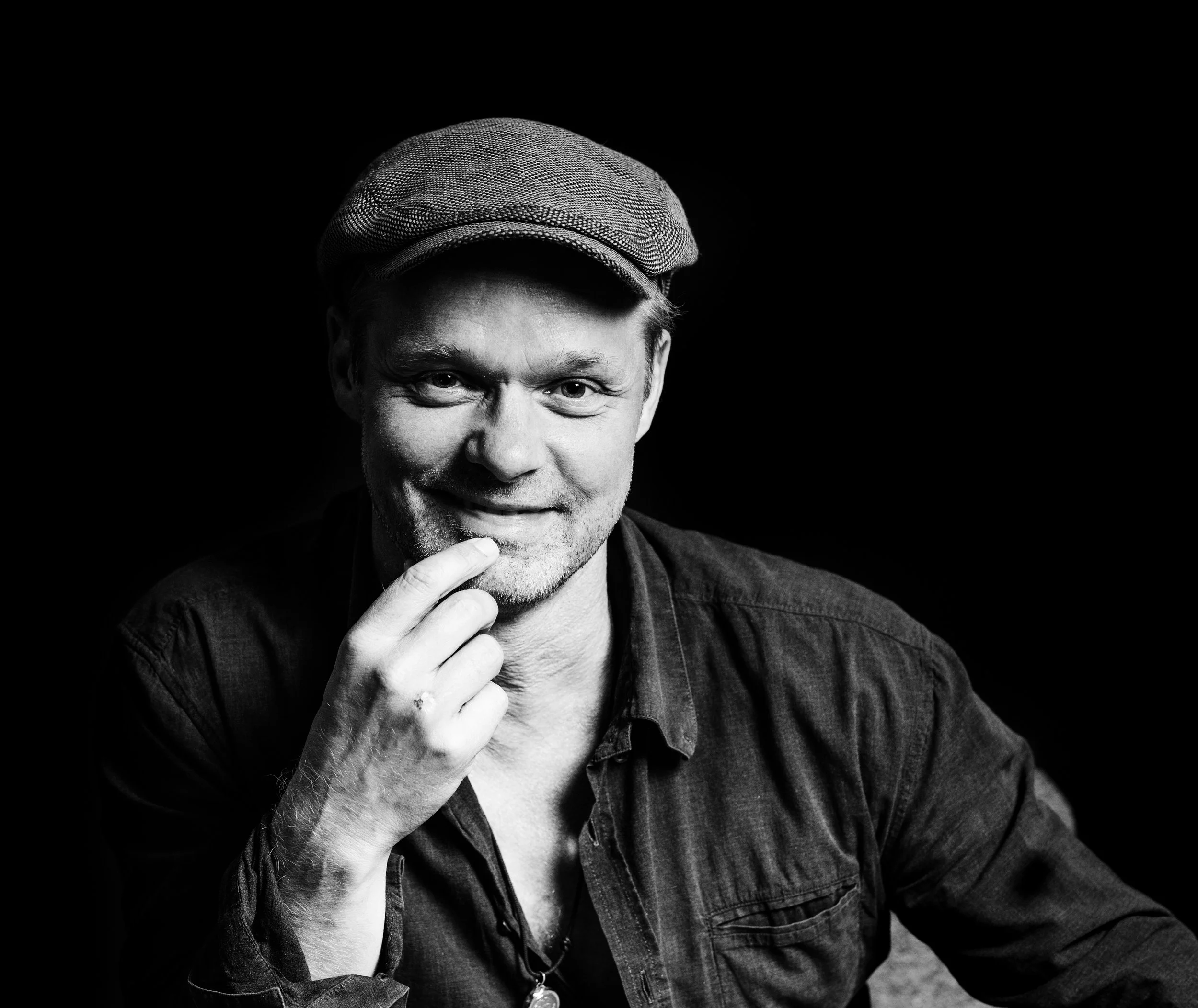 Black and white portrait of a smiling man wearing a cap, with his hand touching his chin, against a dark background.