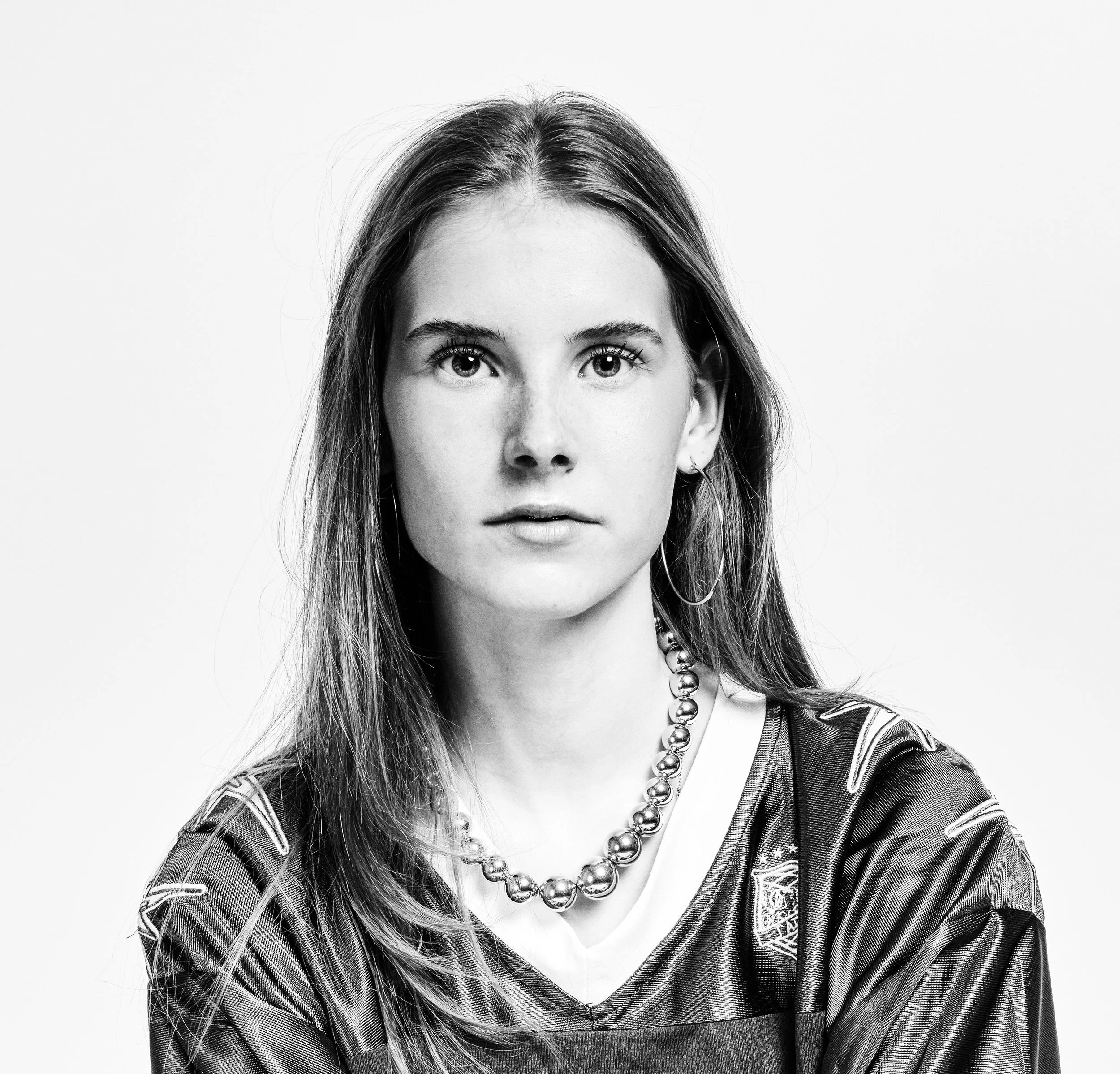 A black and white portrait of a young woman with long hair, wearing a striped shirt, large hoop earrings, and a beaded necklace, looking directly at the camera.