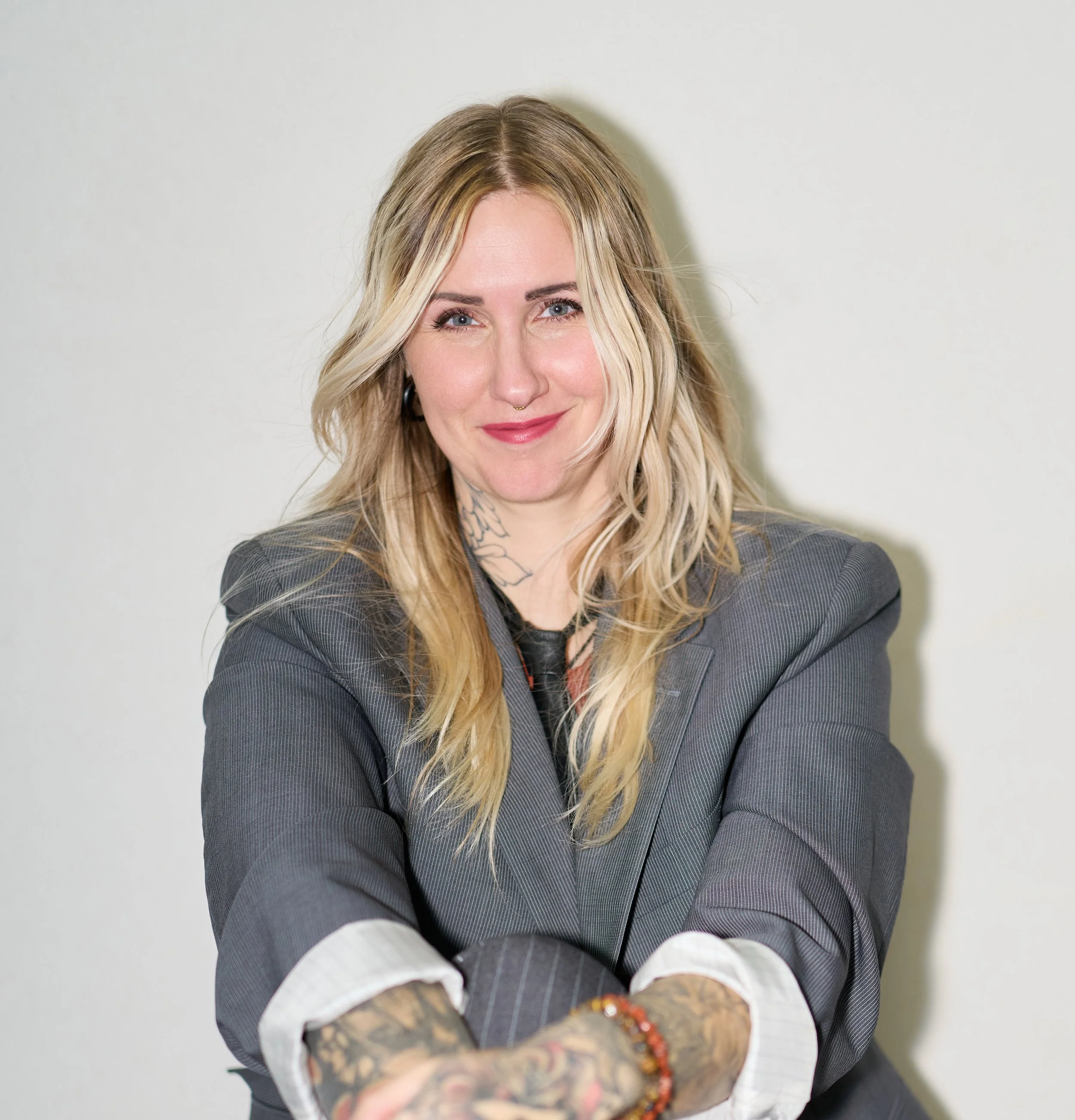 A woman with long blonde hair, wearing a plaid gray blazer and white blouse, smiles at the camera. She has tattoos on her neck and arms, wears a ring and bracelets, and stands against a plain, light-colored wall.