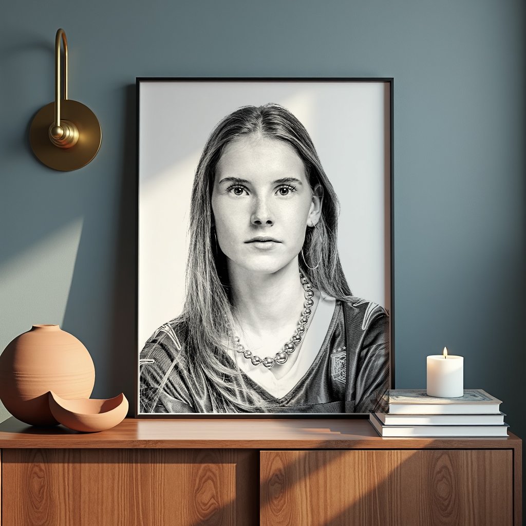 A black and white portrait of a young woman with long hair, wearing a necklace and hoop earrings, displayed in a large frame on a blue wall. A modern gold wall sconce, a ceramic vase with a broken piece, and a lit candle on a stack of books are arranged on a wooden surface below the portrait.