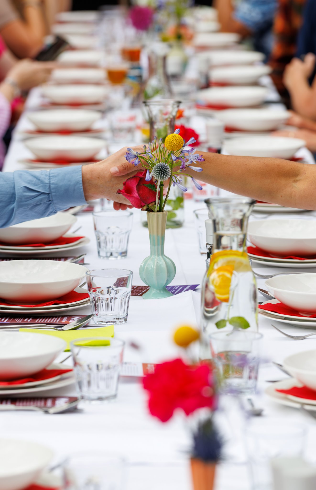 Een tafel gedekt voor een samenkomst, versierd met een vaas bloemen en glazen, met mensen die zich voorbereiden op een maaltijd.