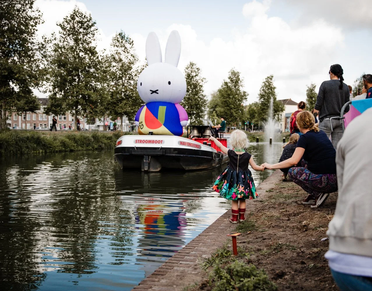 A boat with a large inflatable rabbit figure on top floats on a canal during an outdoor event. Several people, including children, are gathered along the canal's edge, some holding hands and watching the boat.