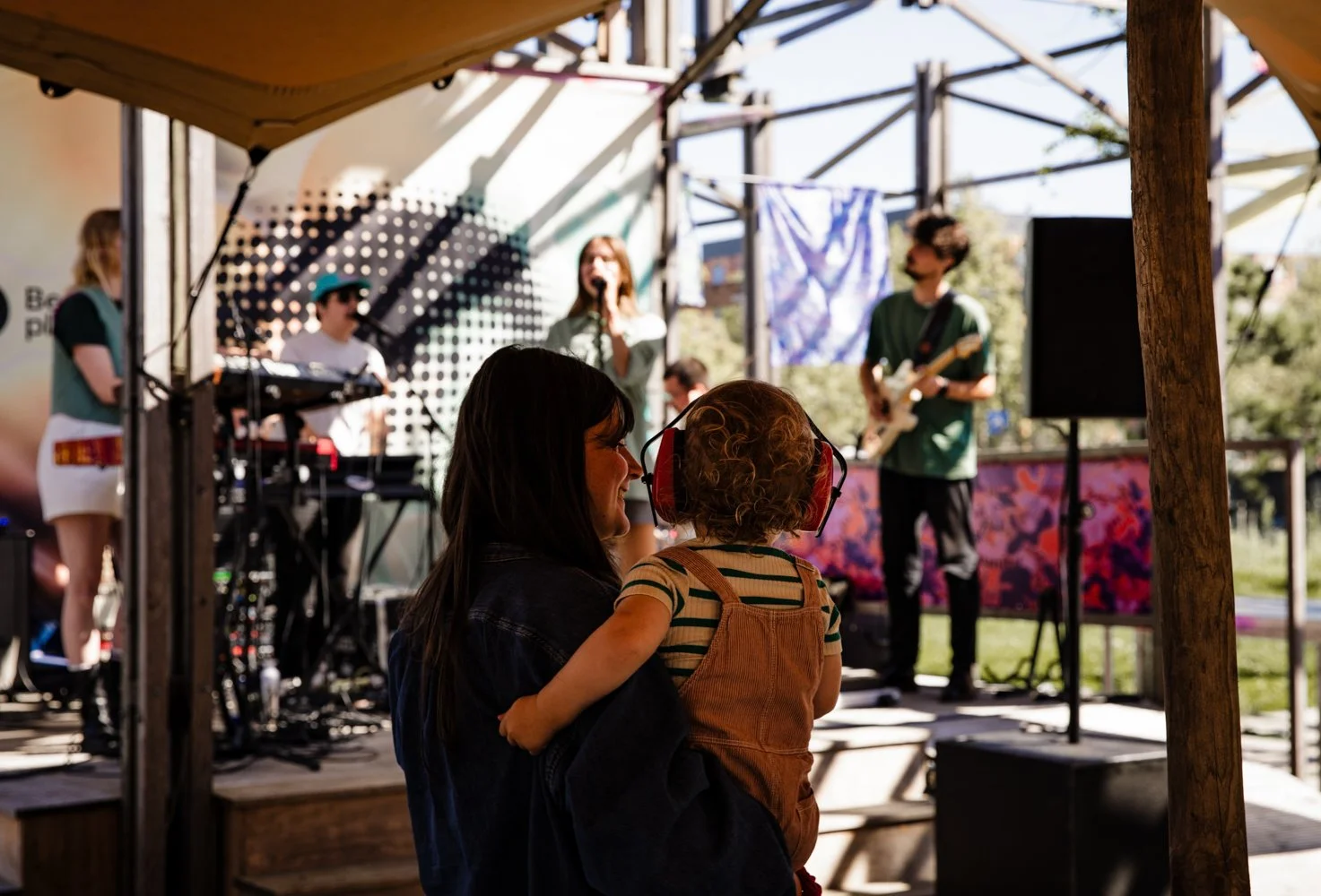 A woman holding a young child wearing headphones, watching a band perform on an outdoor stage under a wooden canopy. The band includes a female singer, a keyboard player, and a guitarist.