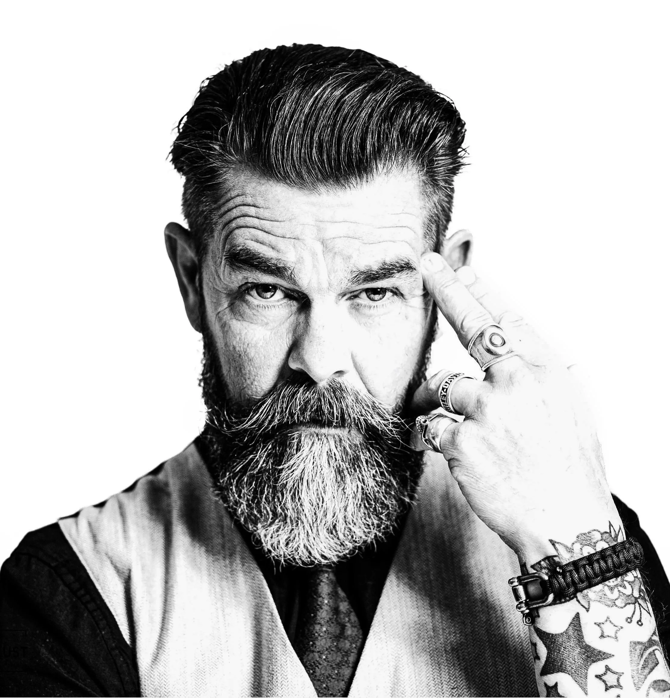 Black and white portrait of a bearded man with styled hair, wearing rings and bracelets, pointing to his temple with his index finger.