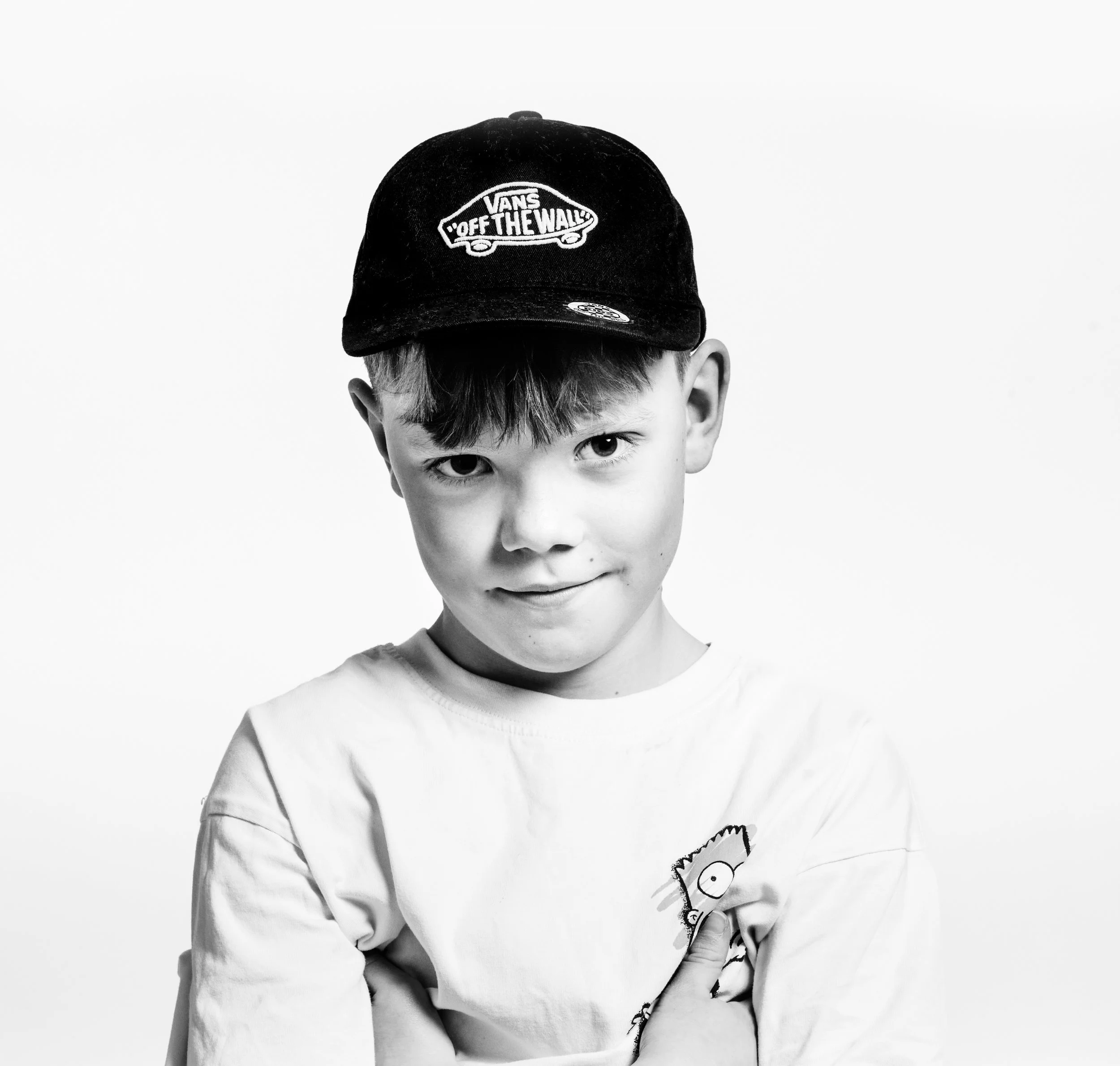 Black and white photo of a young boy with a slight smile, wearing a Vans cap and a T-shirt with a cartoon character on it, arms crossed.