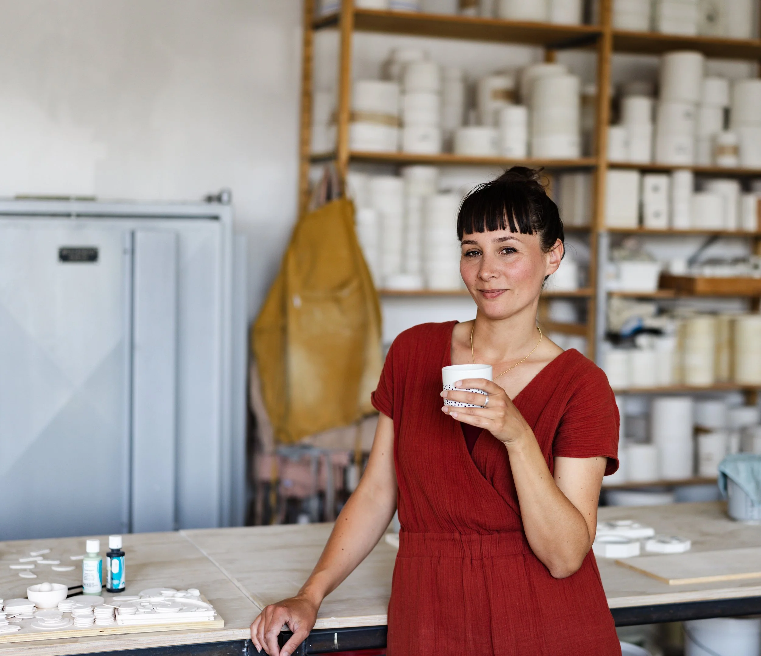 A woman in a red dress holding a white cup, standing in a ceramics studio with shelves of pottery and tools in the background.