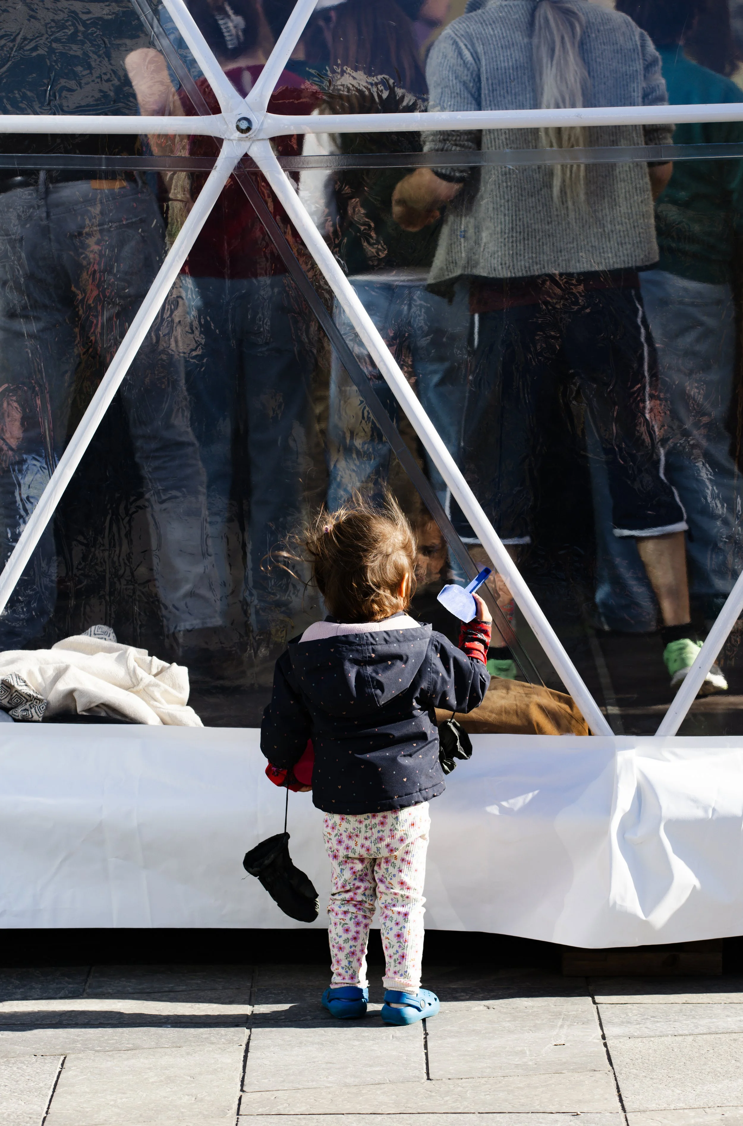 A young girl with curly hair wearing a dark jacket, pink and white floral pants, and blue Crocs, standing on a sidewalk and looking at a group of people through a transparent enclosure, possibly a bus or an exhibit.