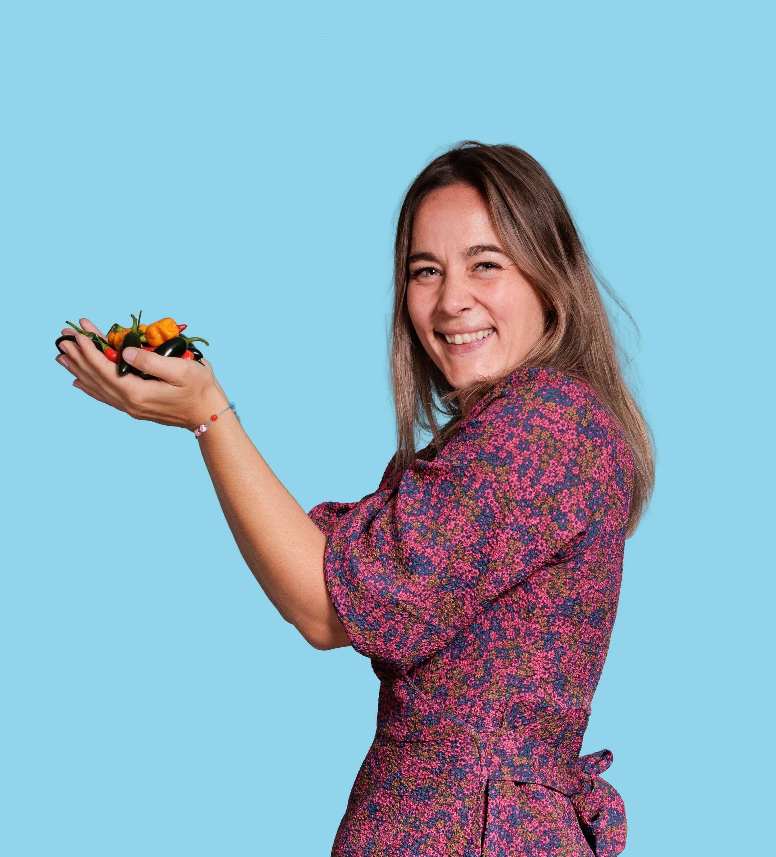 A woman with long light brown hair smiling and holding a bowl of colorful peppers, wearing a colorful patterned dress or top, against a light blue background.