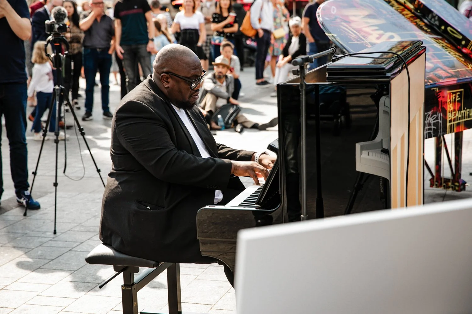 A man wearing glasses and a black jacket plays a piano outdoors while a crowd watches, with some people sitting on the ground and others standing.