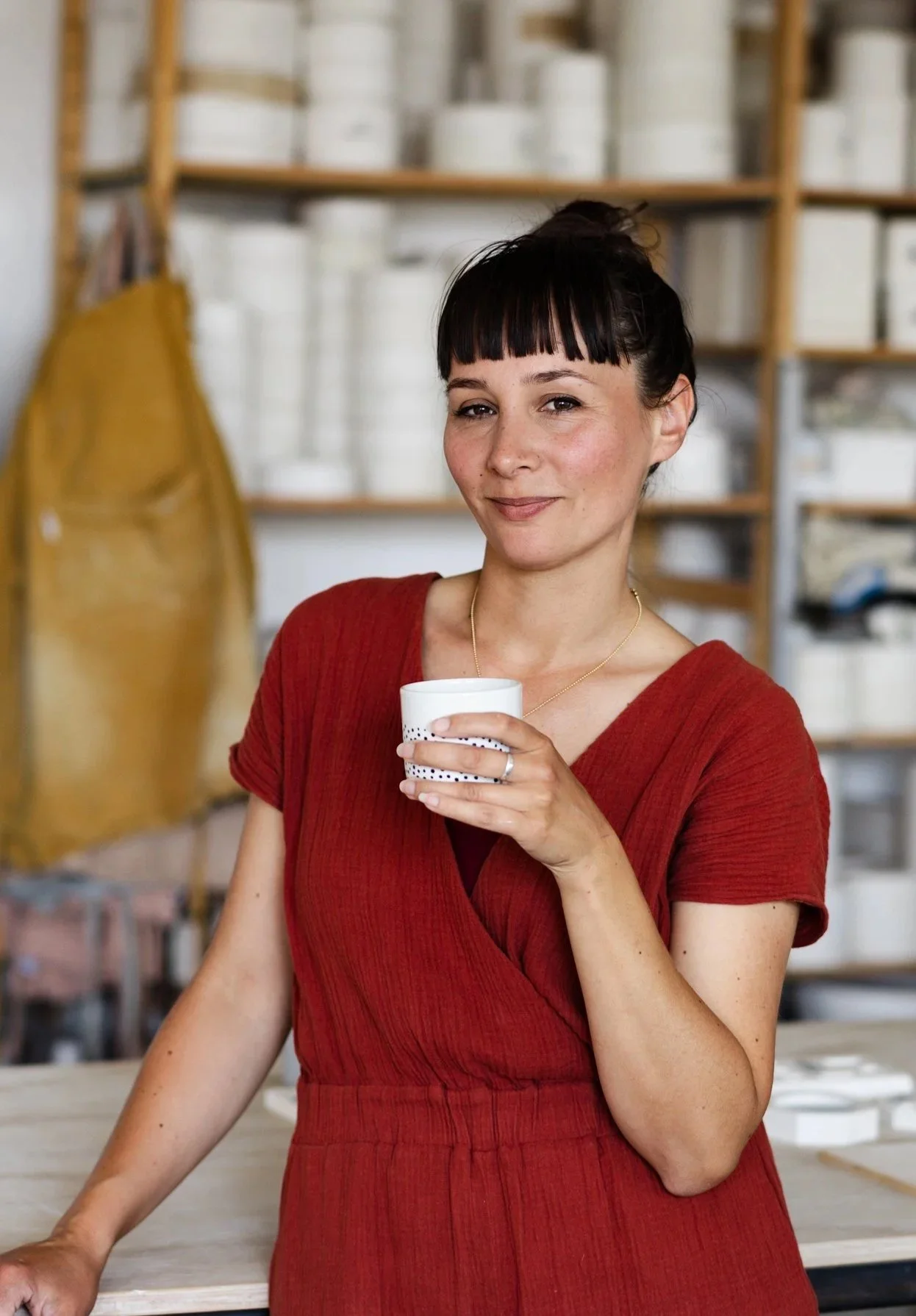 Vrouw met zwart haar en rechte pony, in een kastanjebruine jurk, die een witte beker vasthoudt, binnen in een ruimte met houten planken gevuld met witte dozen.