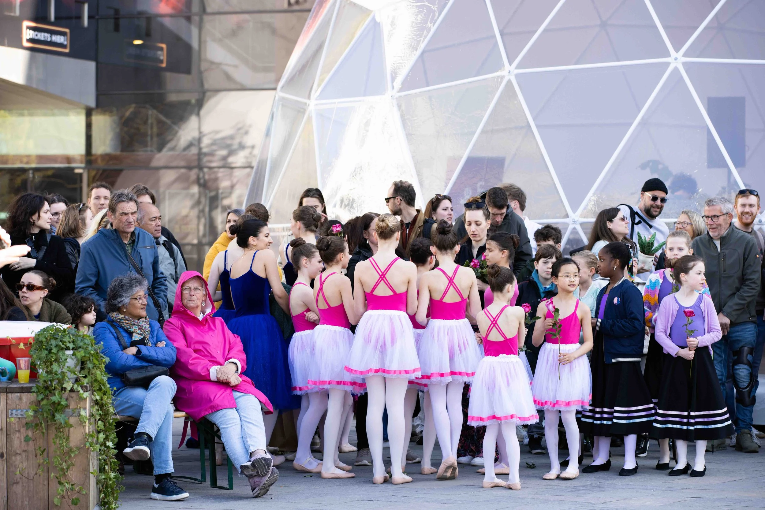 A group of young ballet dancers dressed in pink, purple, and blue costumes are gathered in a circle, holding roses, while adults and seniors observe and take photos in an outdoor urban setting with modern architecture in the background.