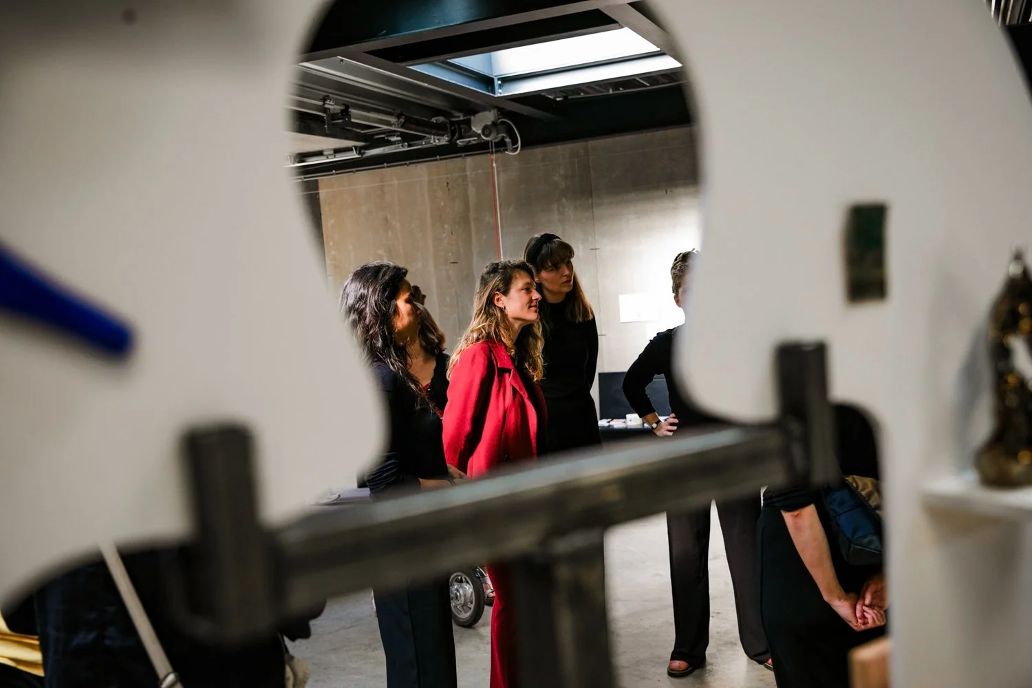 A group of women standing and talking in an industrial-style room, seen through a large, irregularly shaped cutout in a piece of equipment or machinery.
