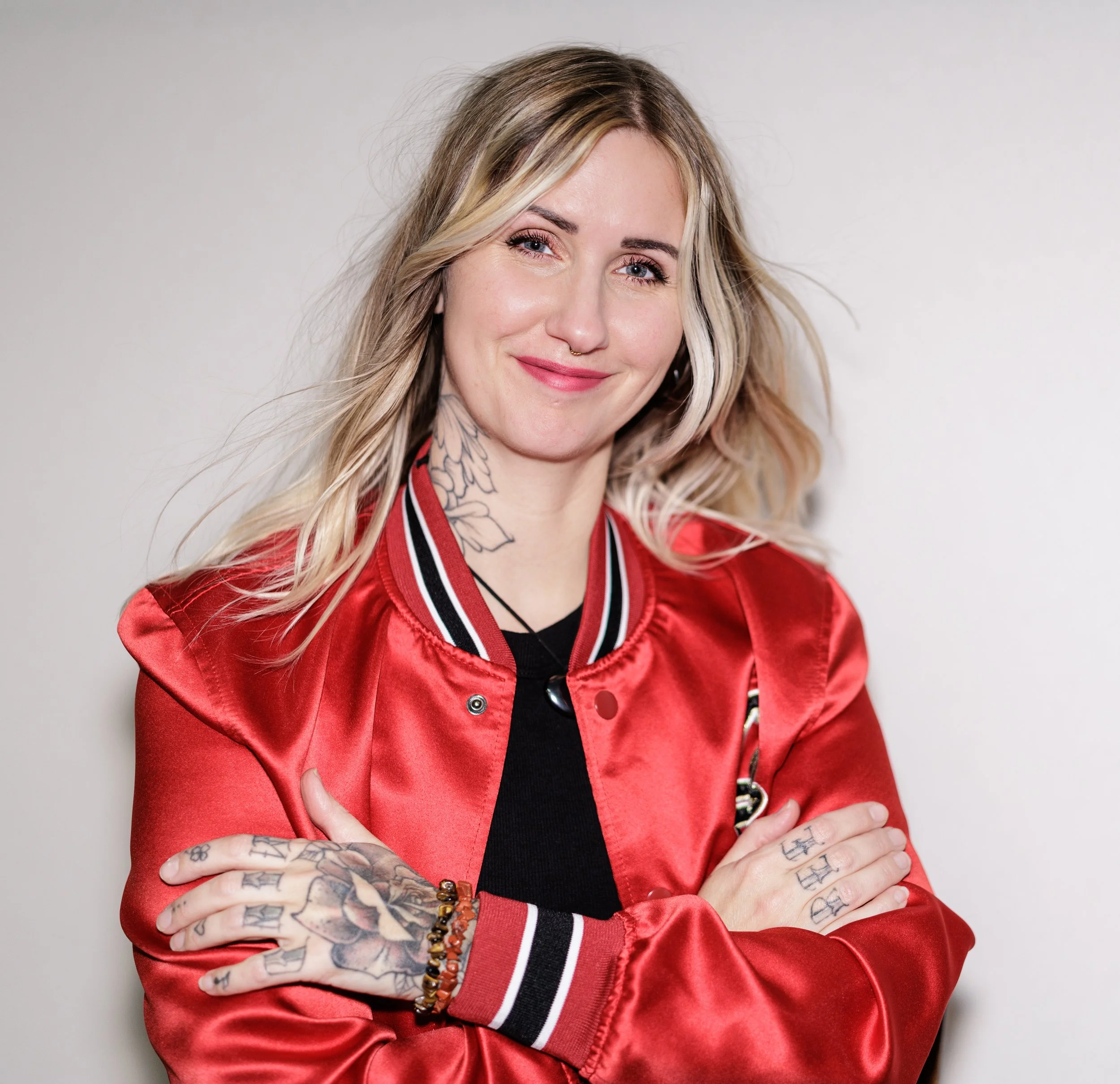 A young woman with blonde hair, tattoos on her hands and neck, wearing a red satin jacket with black and white accents, smiling with her arms crossed against a plain background.