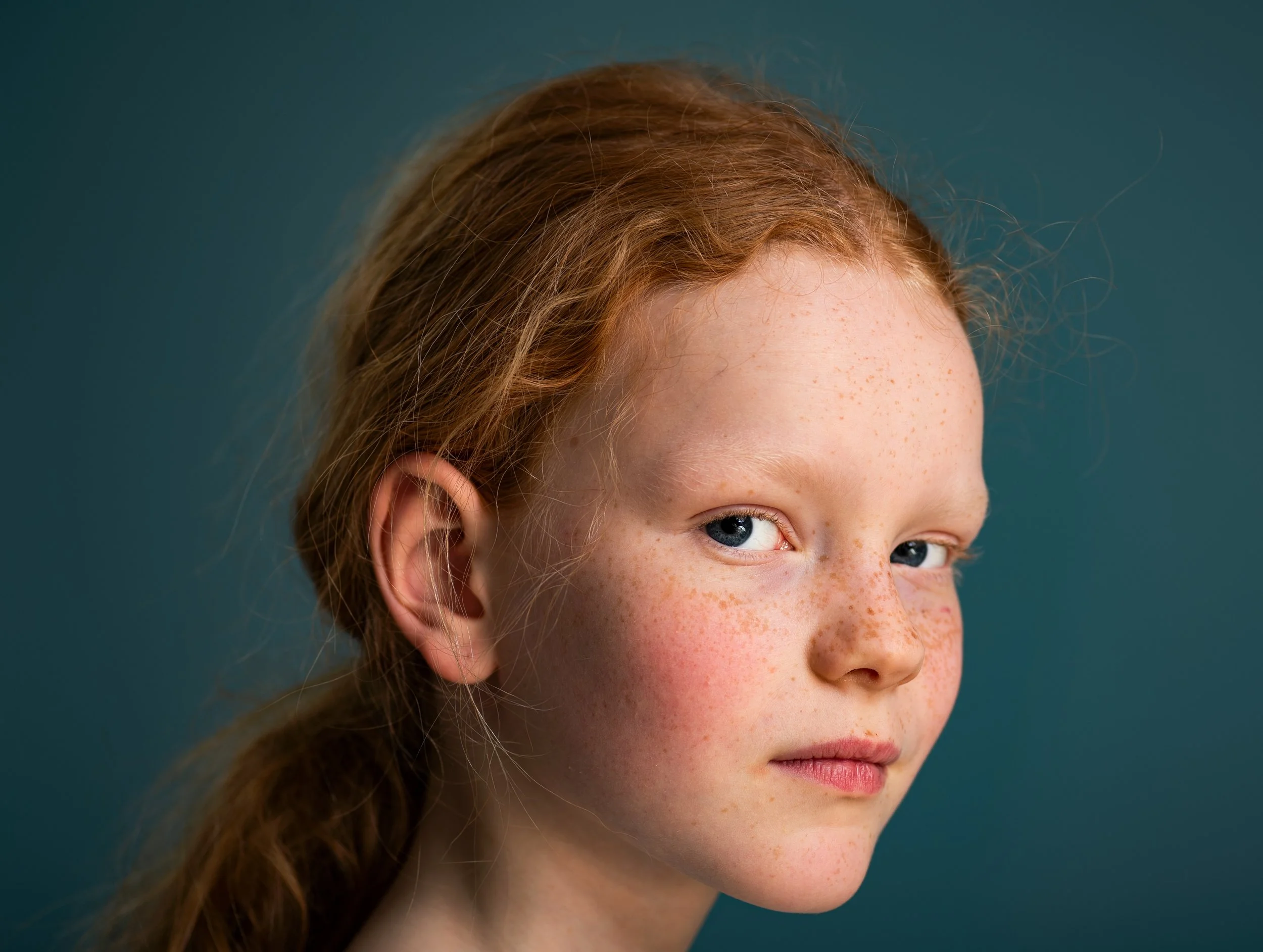 A close-up portrait of a young girl with red hair, freckles, and blue eyes, looking slightly to the side against a dark teal background.
