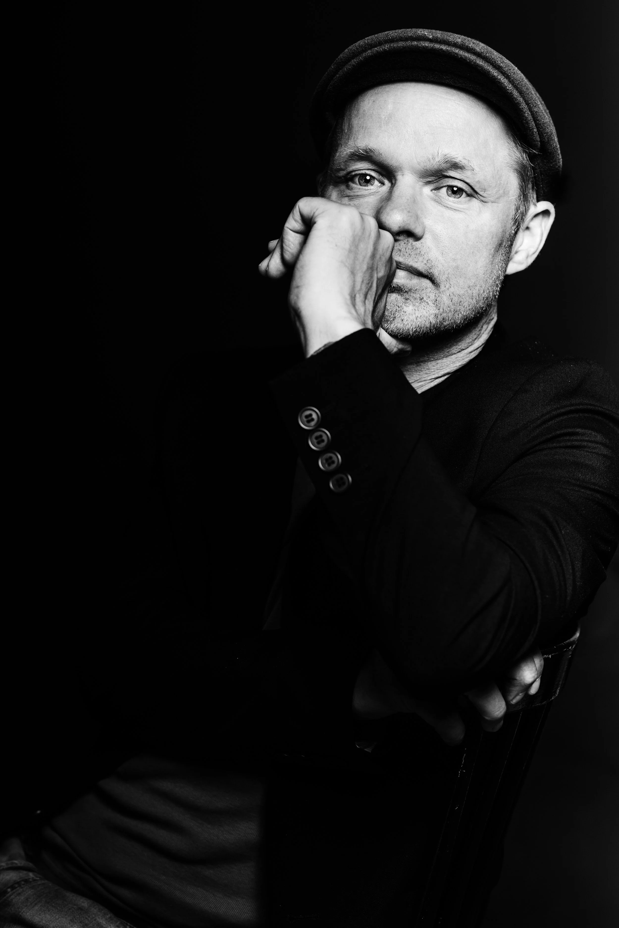 Black and white portrait of a man wearing a hat, sitting with his hand resting on his face, looking contemplative.