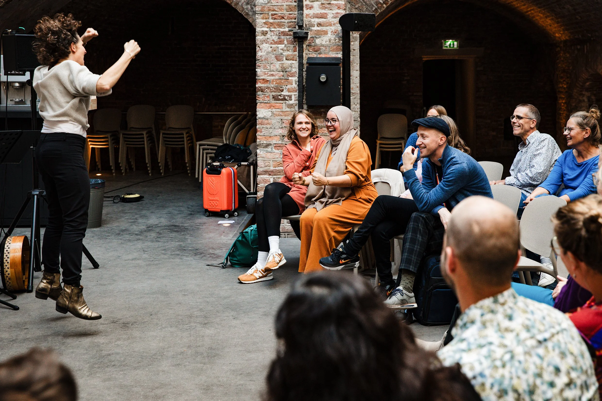 A woman with curly hair, wearing a beige sweater and black pants, is performing on stage in front of an audience. The audience is seated, laughing, and appears to be enjoying the performance, in a venue with brick walls and an arched ceiling.