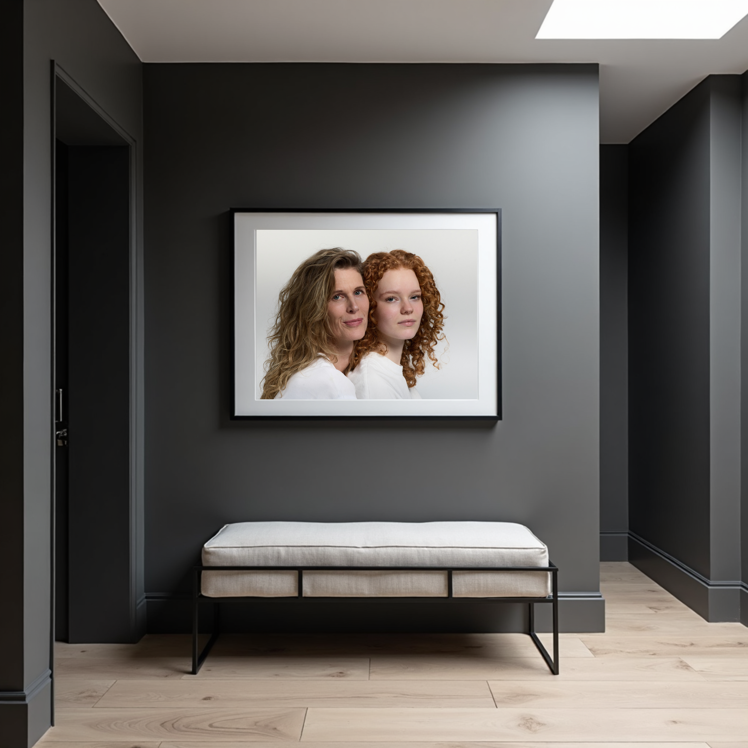A framed portrait of two women with curly hair, one with light brown and one with red hair, hangs on a gray wall in a modern, minimalist hallway. A beige cushioned bench with a black metal frame is positioned below the artwork.