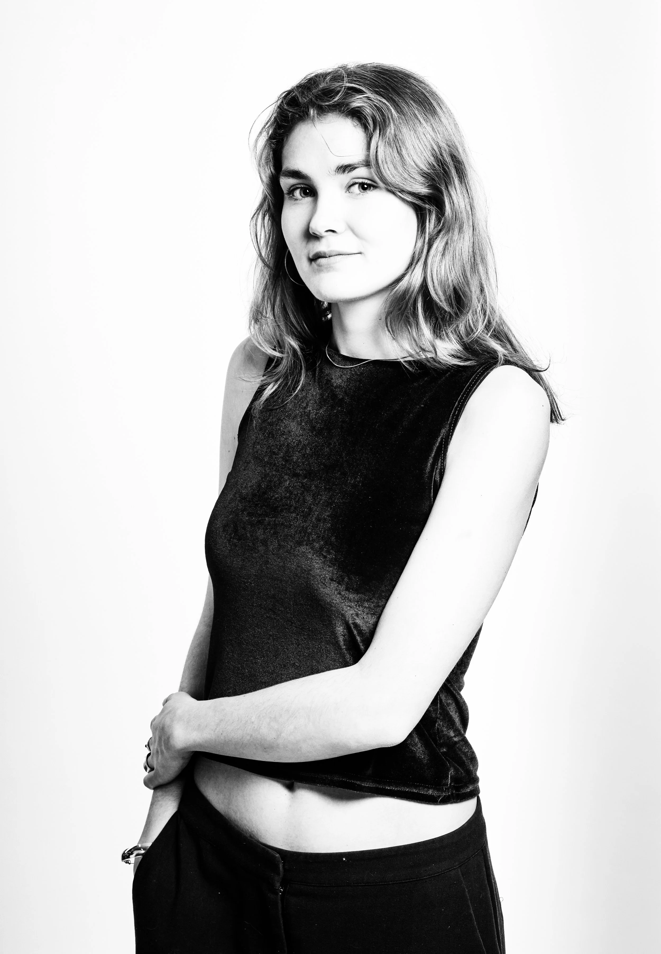 Black and white portrait of a young woman with shoulder-length hair, wearing a sleeveless velvet top and black pants, standing against a plain background.