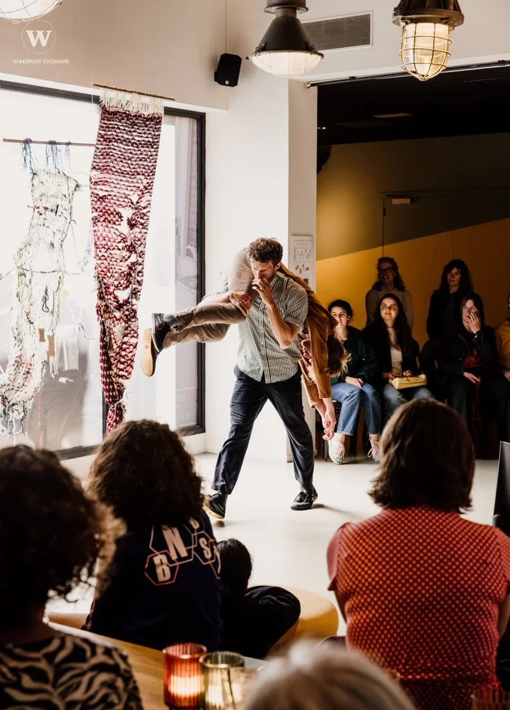 A man is balancing a child on his shoulders in a room with a seated audience. The audience appears engaged, watching the performance. The room has modern lighting, a large window with curtains, and artwork or decorations on the wall.