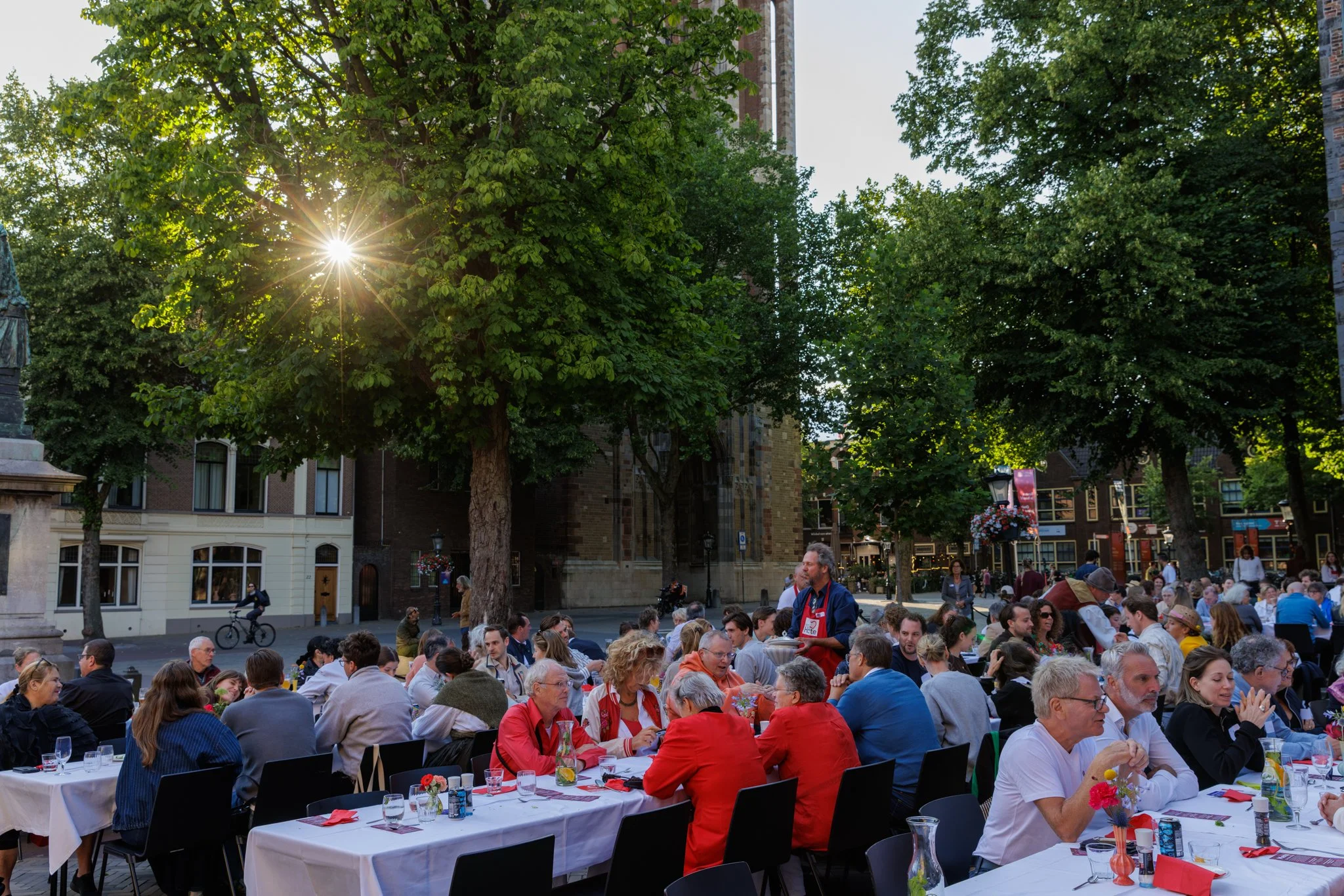 Een druk evenement buiten tijdens de avond met veel mensen zittend aan lange tafels, omringd door bomen en historische gebouwen.