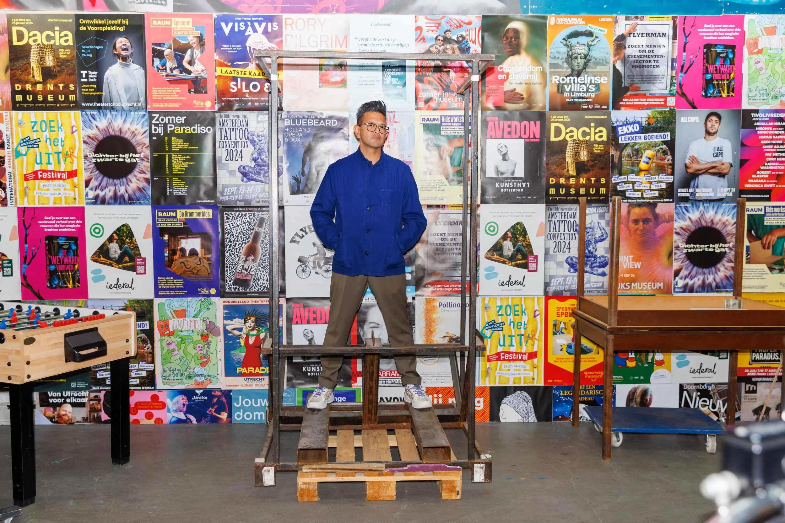 A man wearing glasses, a blue jacket, and beige pants standing on a wooden platform in front of a colorful wall covered with various posters and advertisements, inside an indoor space that appears to be a game or activity center.
