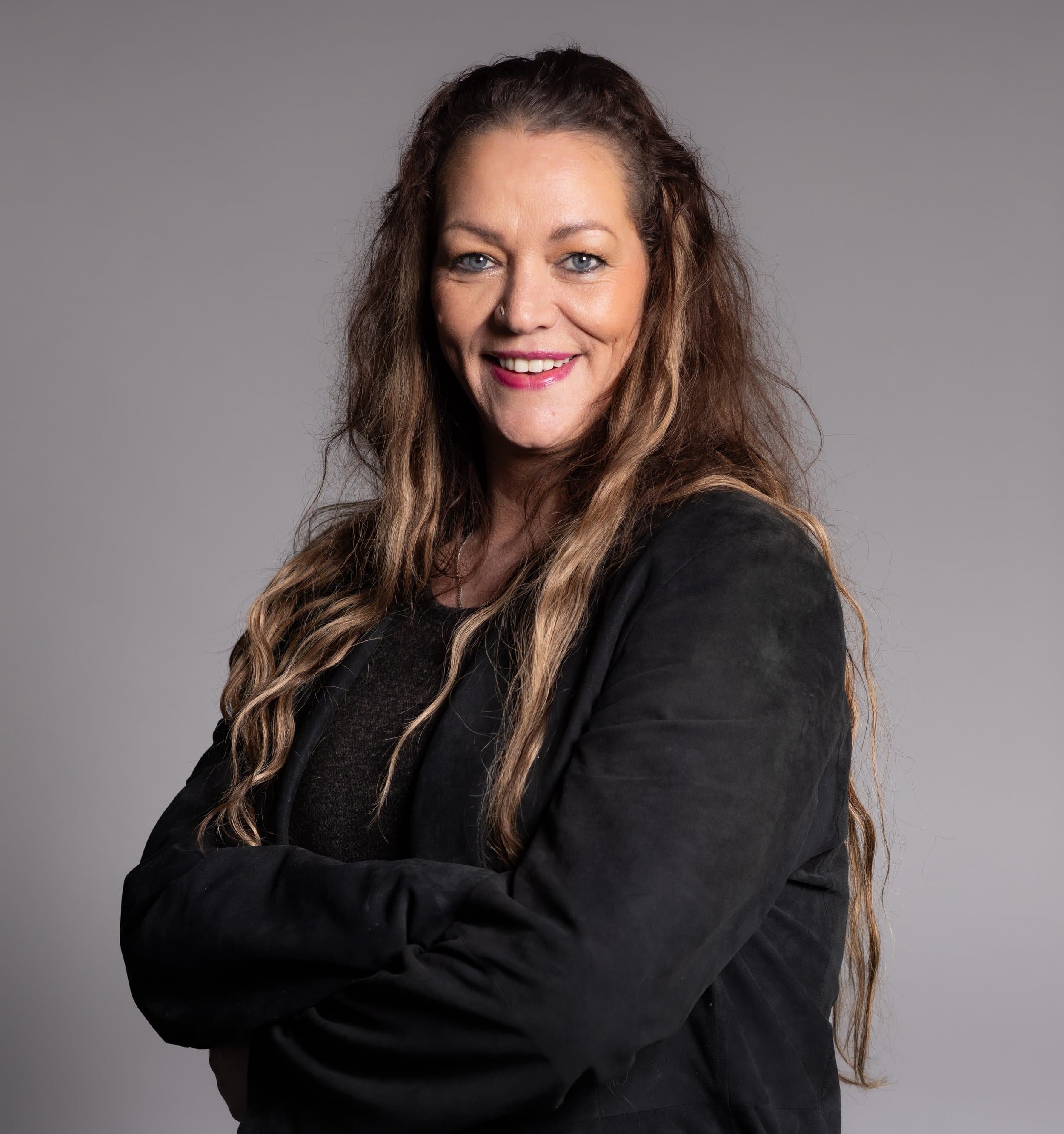 A woman with long, wavy brown hair, blue eyes, and pink lipstick smiling, wearing a black jacket with arms crossed, against a light gray background.
