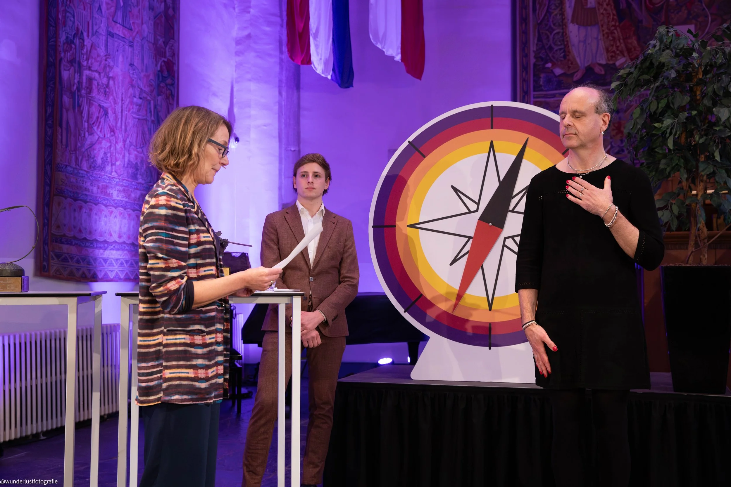 Three people on stage during a ceremony, one woman reading from paper, one man with hand over heart, a young man observing in the background, large compass decoration, colorful banners hanging above, purple lighting, ornate tapestries on the wall.