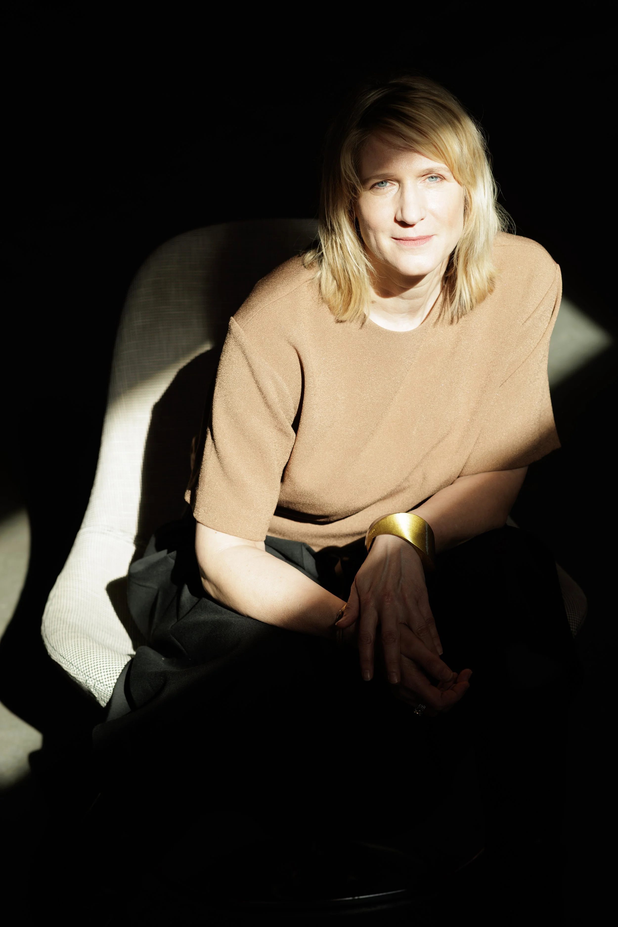 A woman with blonde hair and blue eyes sitting in a dark room, illuminated by light, wearing a beige top and a gold bracelet.