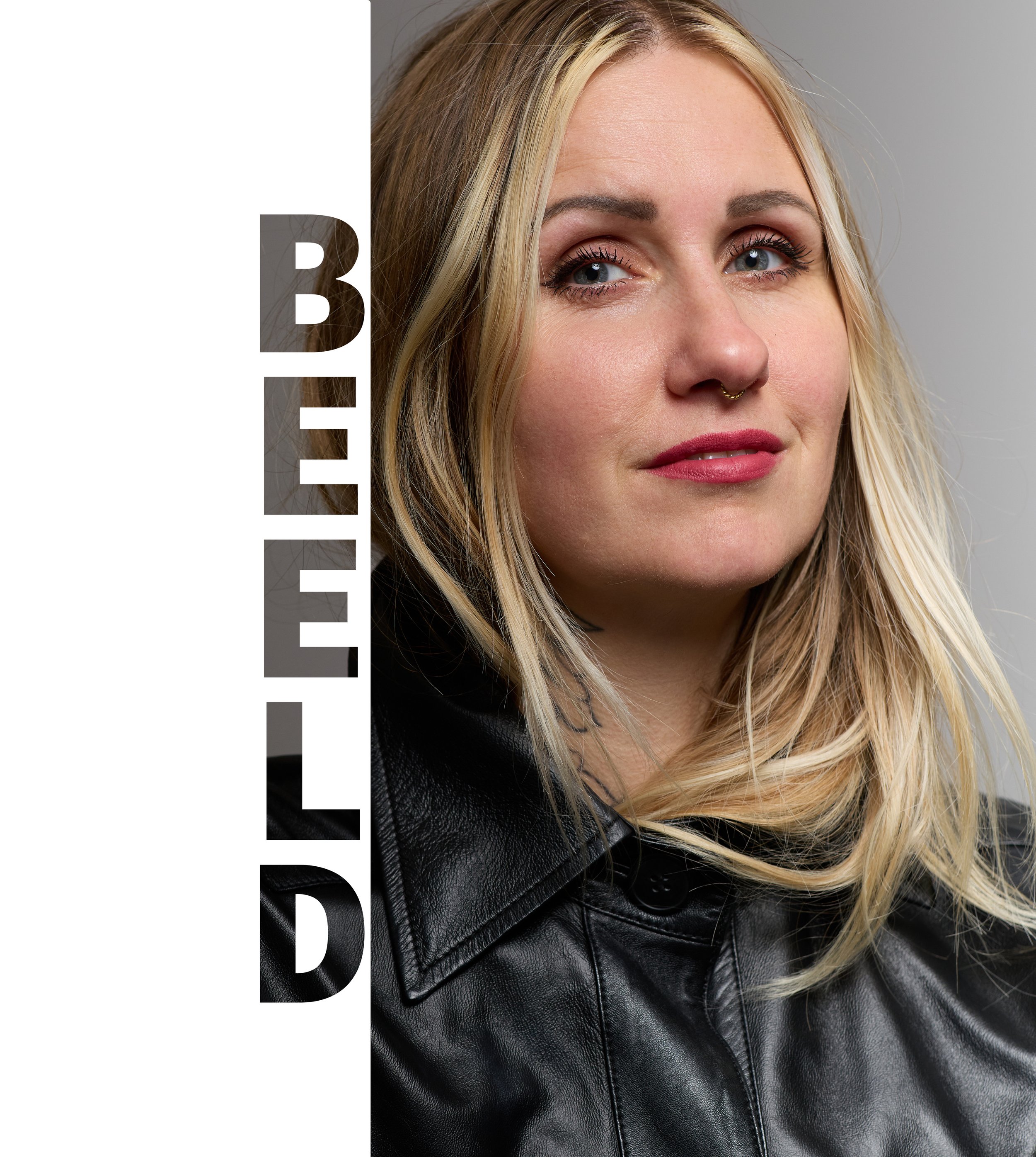 Close-up van een vrouw met blond haar, blauwe ogen, een neusring en een zwarte leren jas, zittend op een witte achtergrond. Het woord 'BEER' staat verticaal over de linkerhelft van de afbeelding.