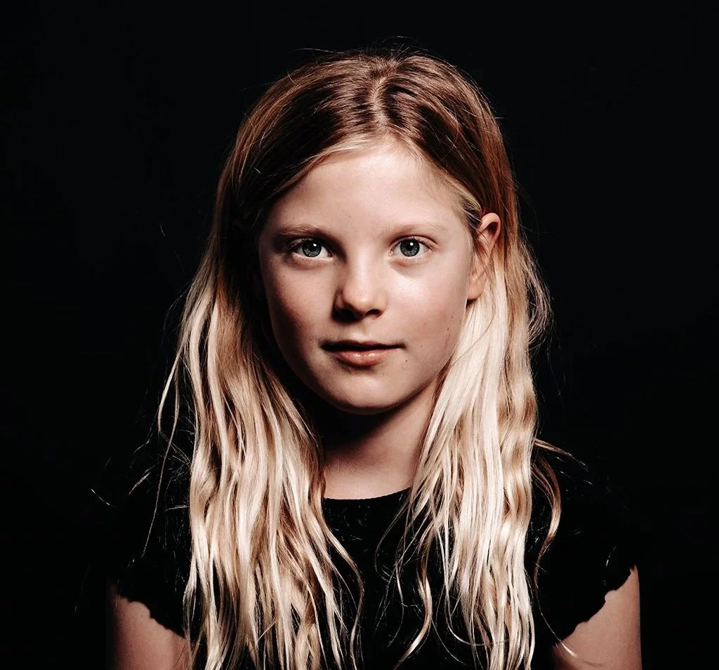 Portret van een jonge vrouw met lange, blonde, golvende haar, kijkend naar de camera met een lichte glimlach, tegen een zwarte achtergrond.
