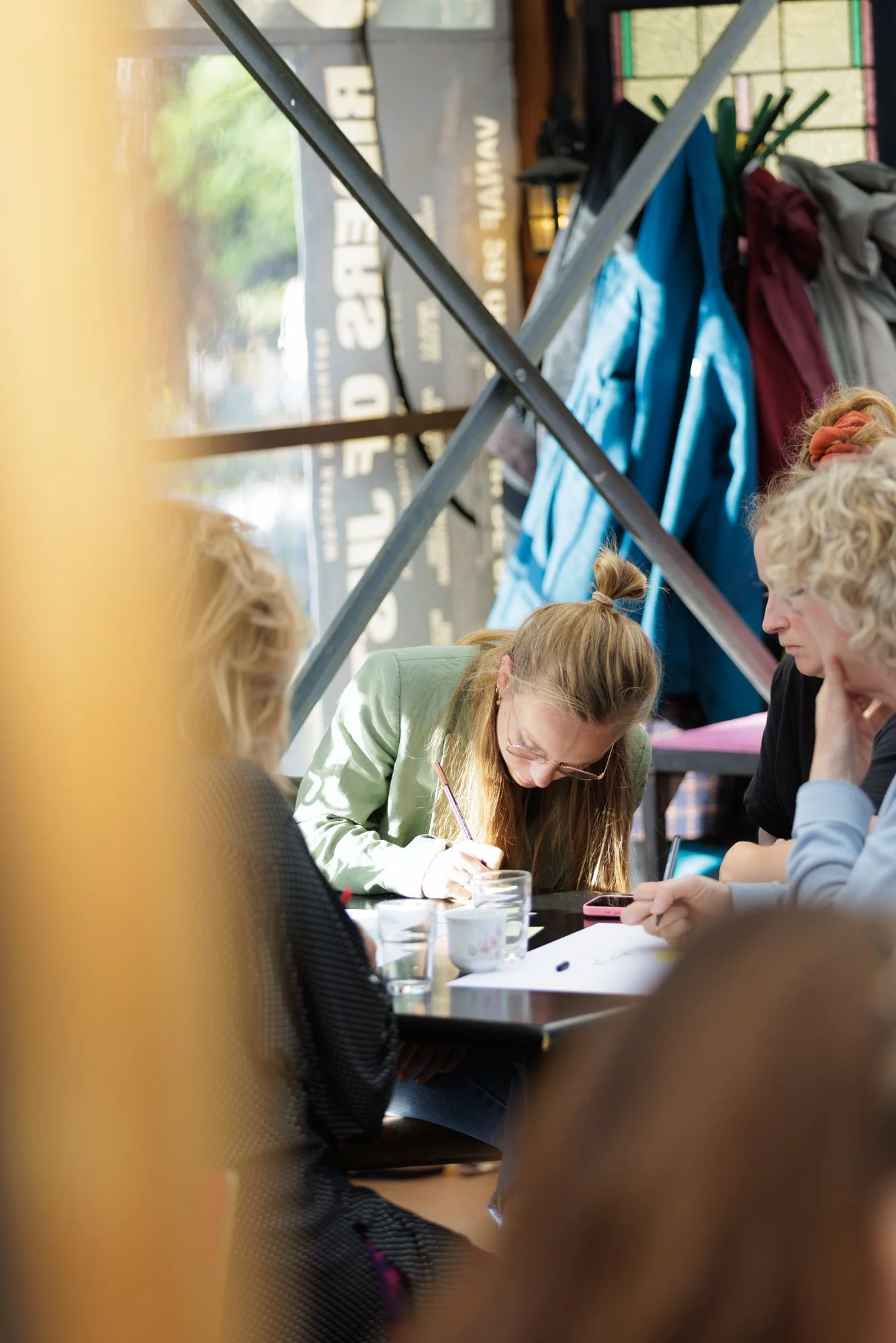 People sitting at a table engaged in a discussion or activity, with various items like notebooks, pens, glasses, and phones on the table, and jackets hanging in the background.